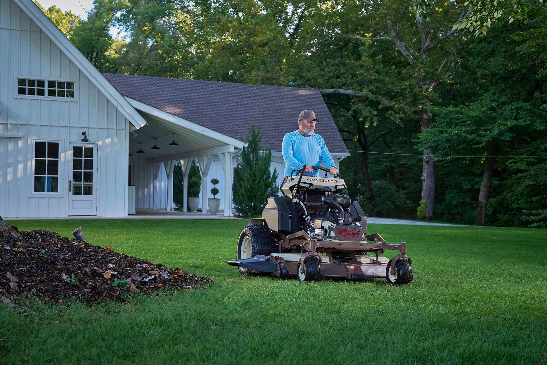 Operator on a Grasshopper stand-on mower cutting lawn beside a mulch pile and white barn.