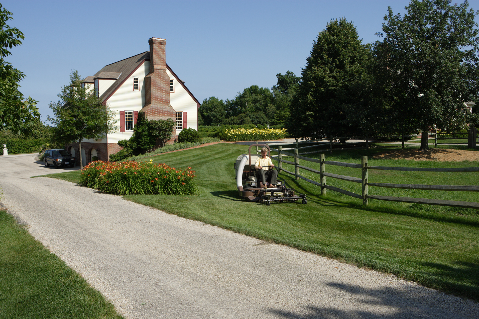 Operator mowing a striped lawn beside a split-rail fence, with a white colonial house behind.