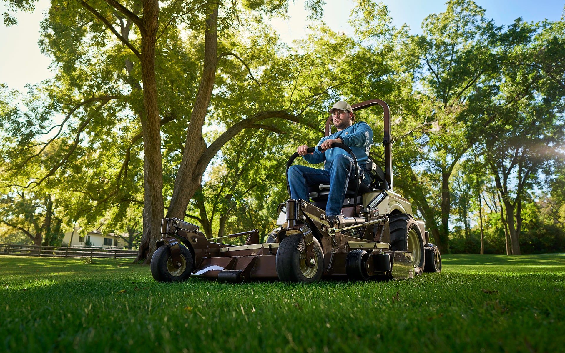 Operator on a Grasshopper FrontMount mower cutting lush lawn beneath tall shade trees.