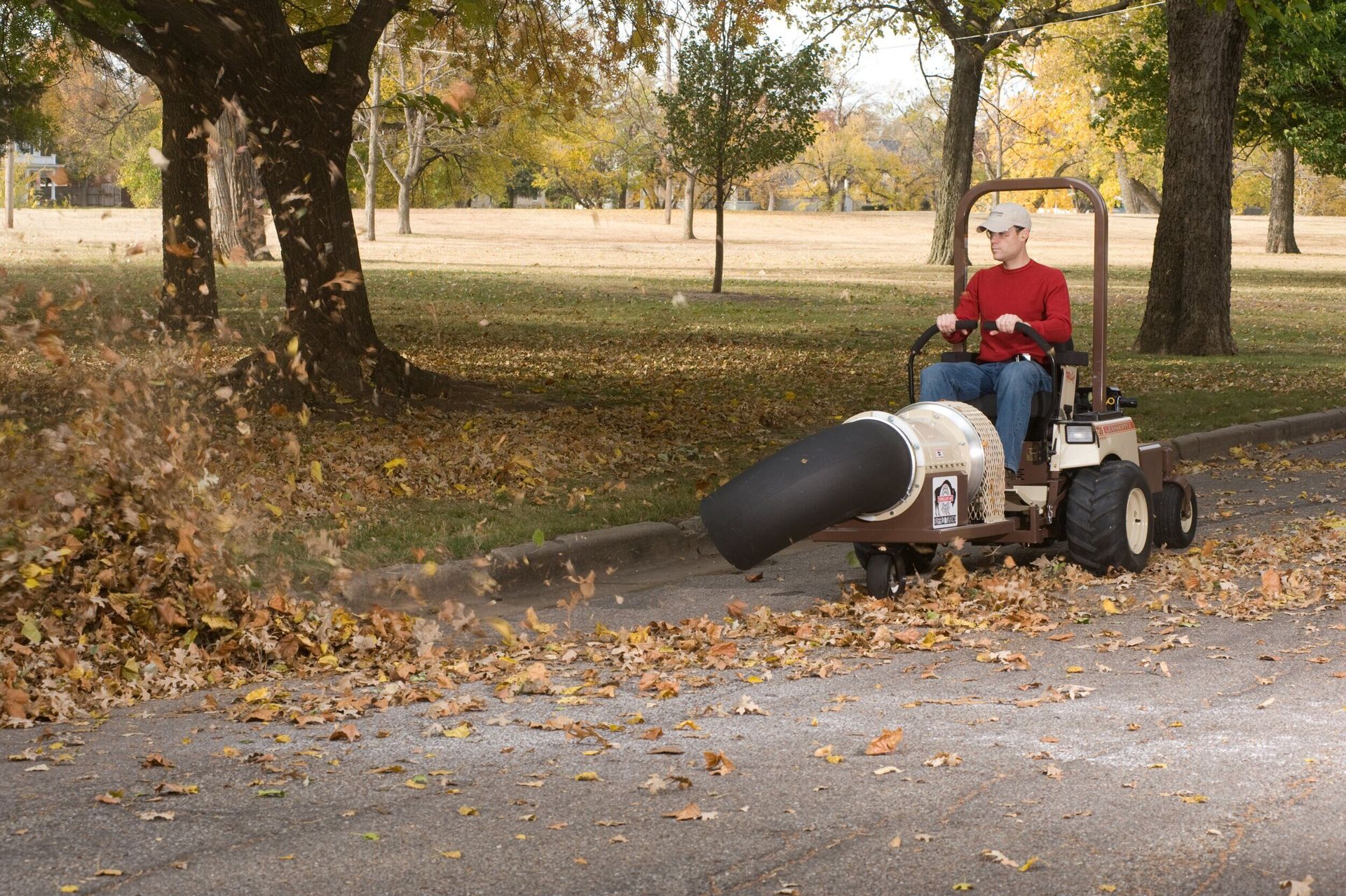 Man riding a Grasshopper mower with a blower attachment, clearing fallen leaves along a park street.