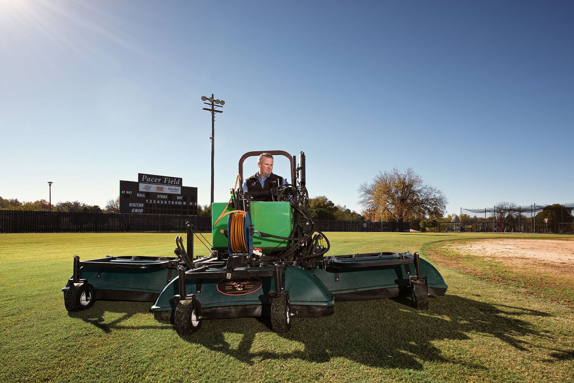 Wide view of a green shielded sprayer rig with three spray hoods parked on ballfield turf.