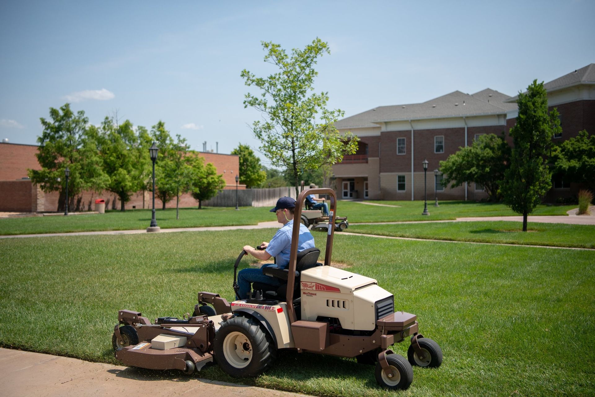 Operator mowing a campus lawn on a Grasshopper 900D diesel FrontMount mower, second machine behind.