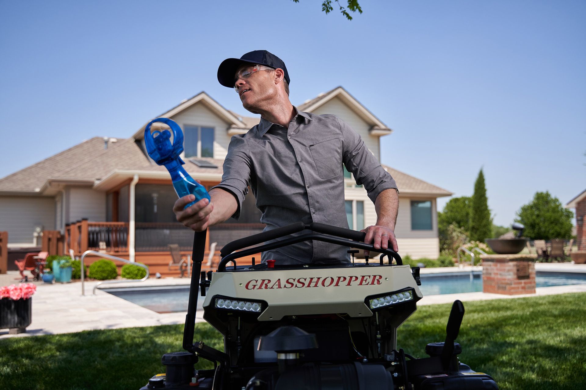 Operator holding a blue misting fan while standing at a Grasshopper mower beside a backyard pool.