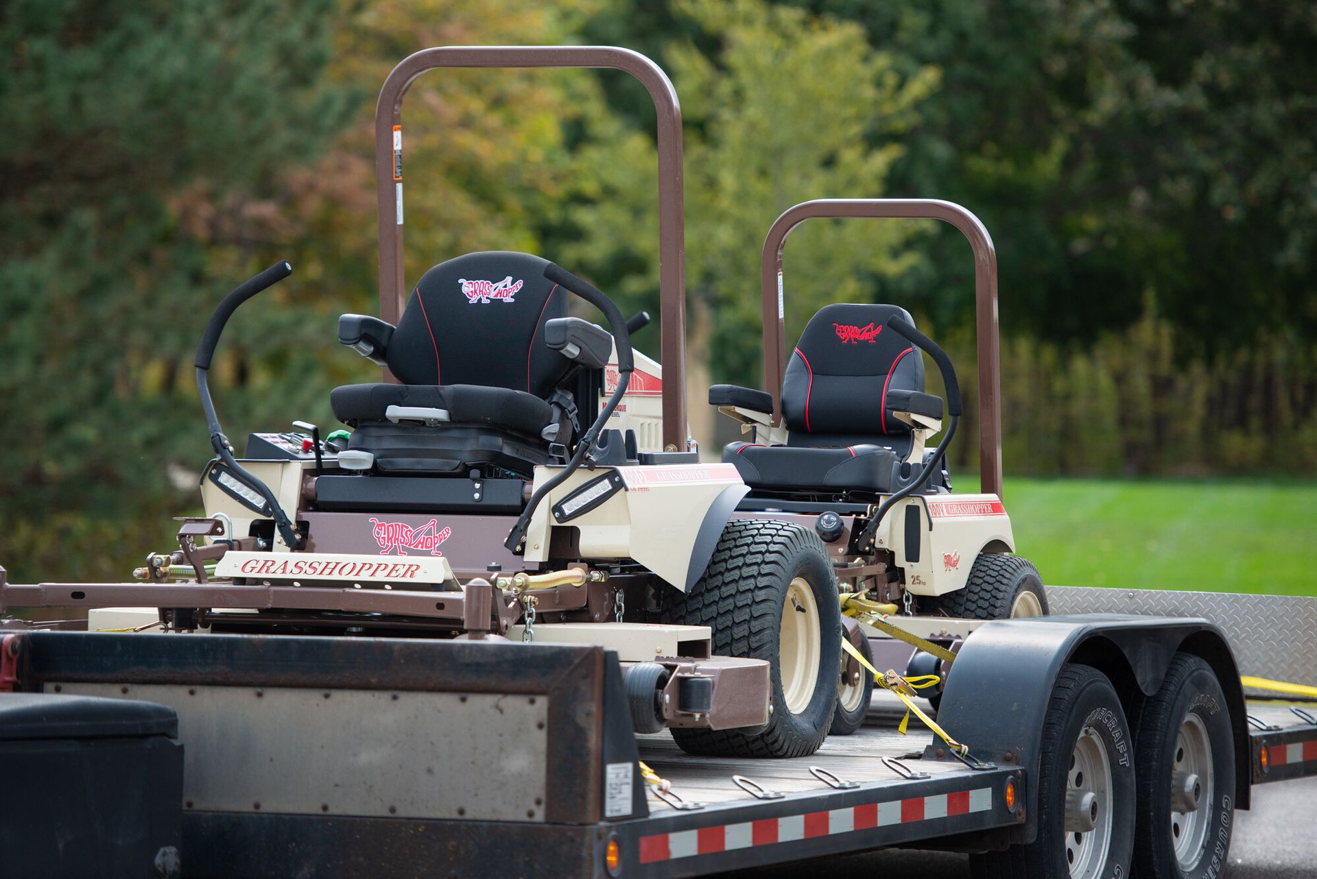 Two Grasshopper mowers, including a 100V, strapped side by side on a flatbed equipment trailer.