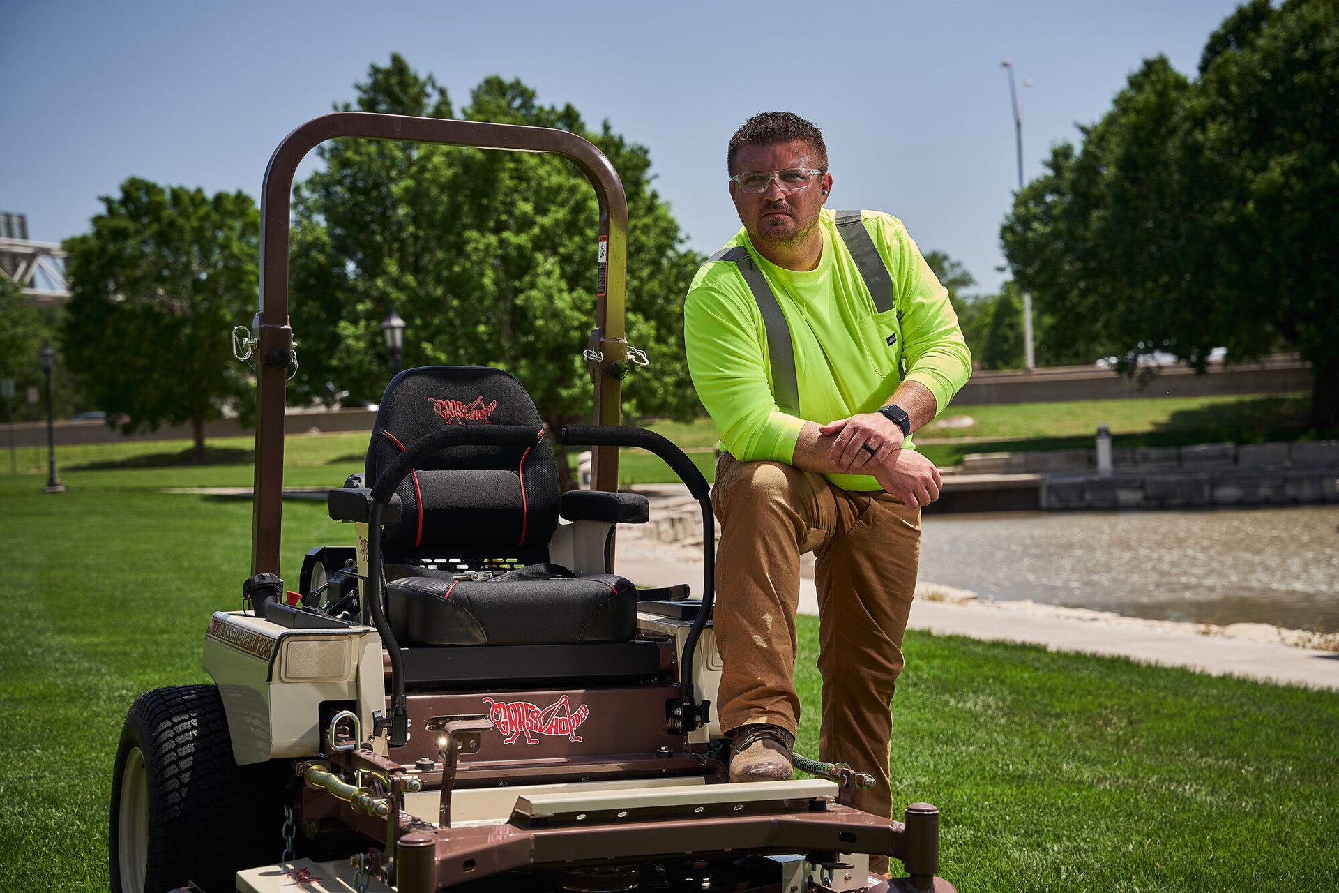Grounds worker in a hi-vis shirt and safety glasses leaning on a Grasshopper mower's rollbar.