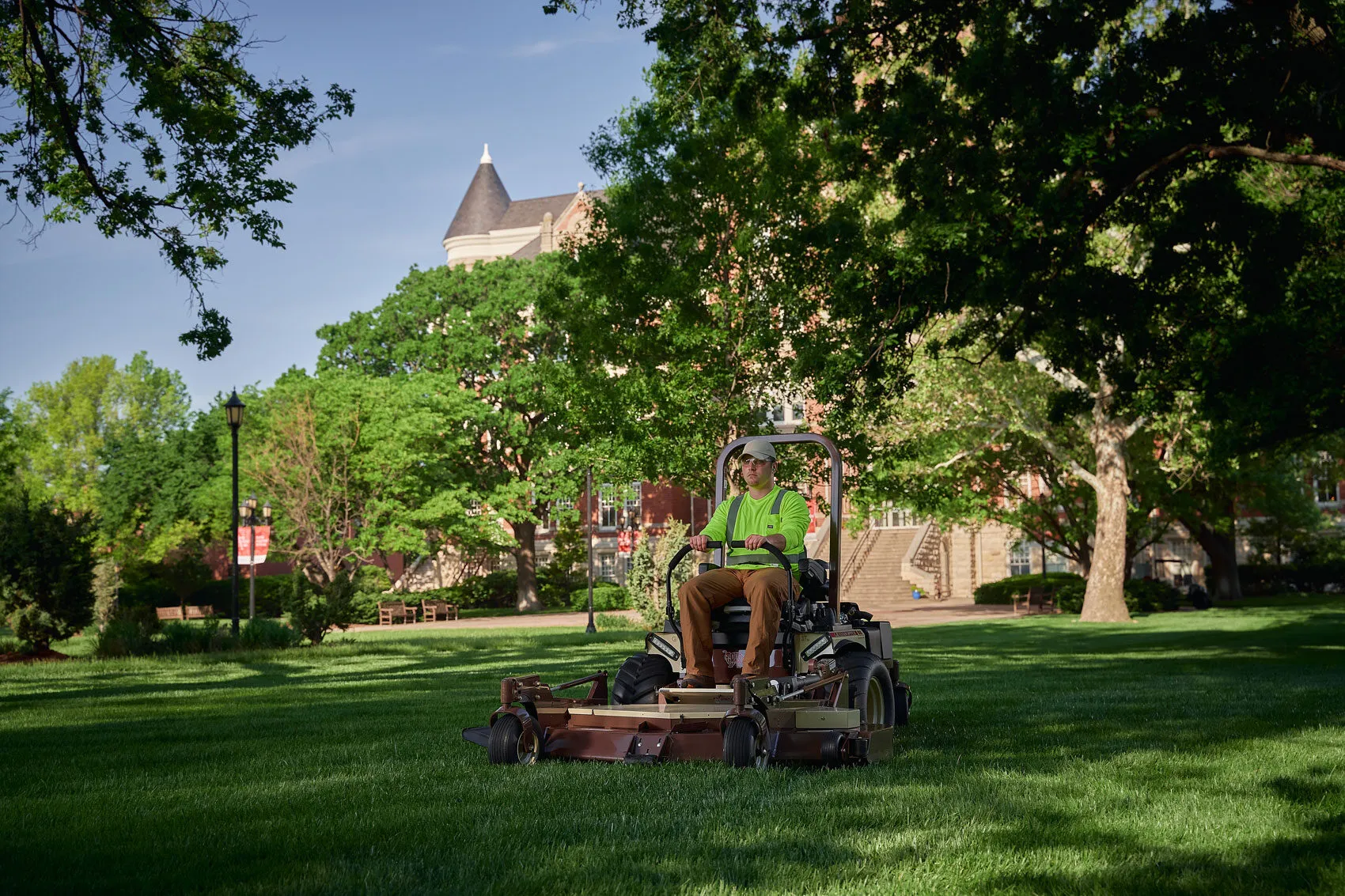 Operator in a hi-vis shirt mowing a shaded campus lawn on a Grasshopper FrontMount mower.