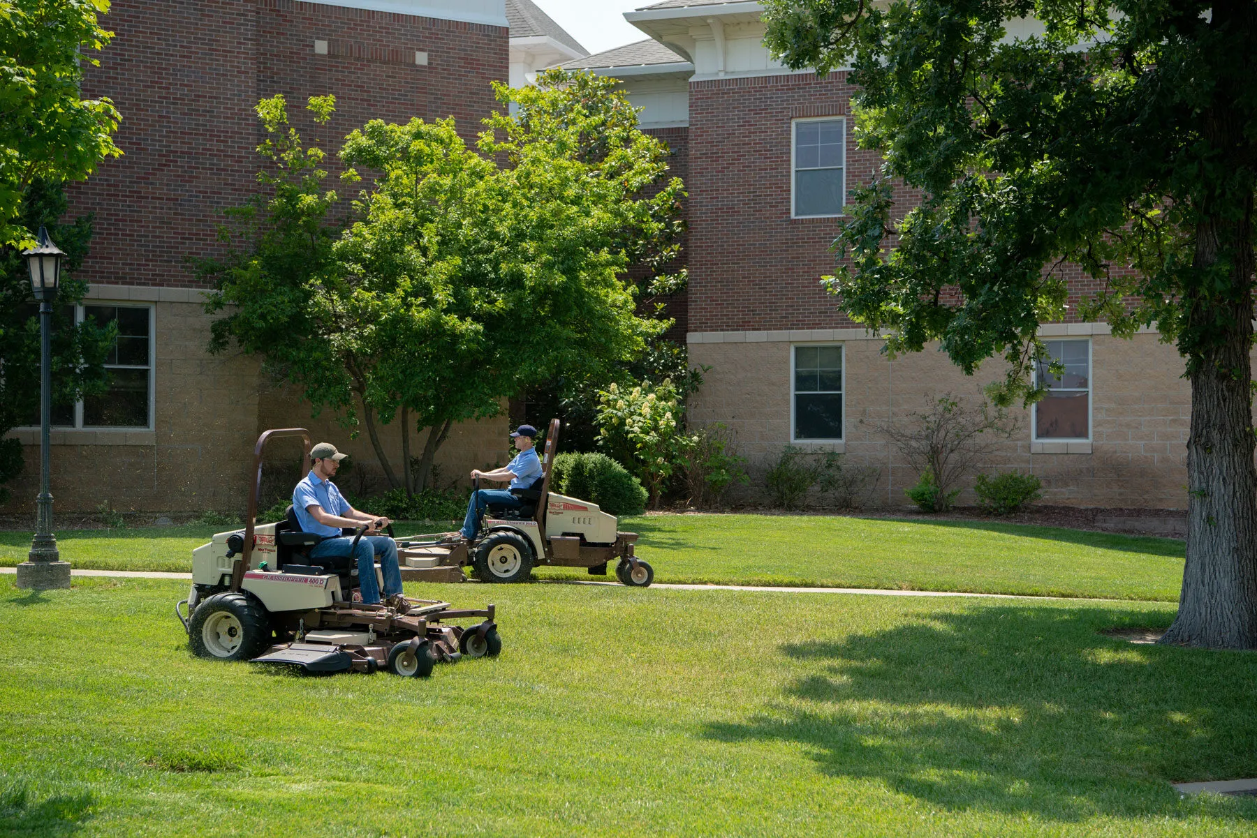 Two operators mowing a shaded campus lawn on Grasshopper FrontMount mowers, the nearest a 400D.