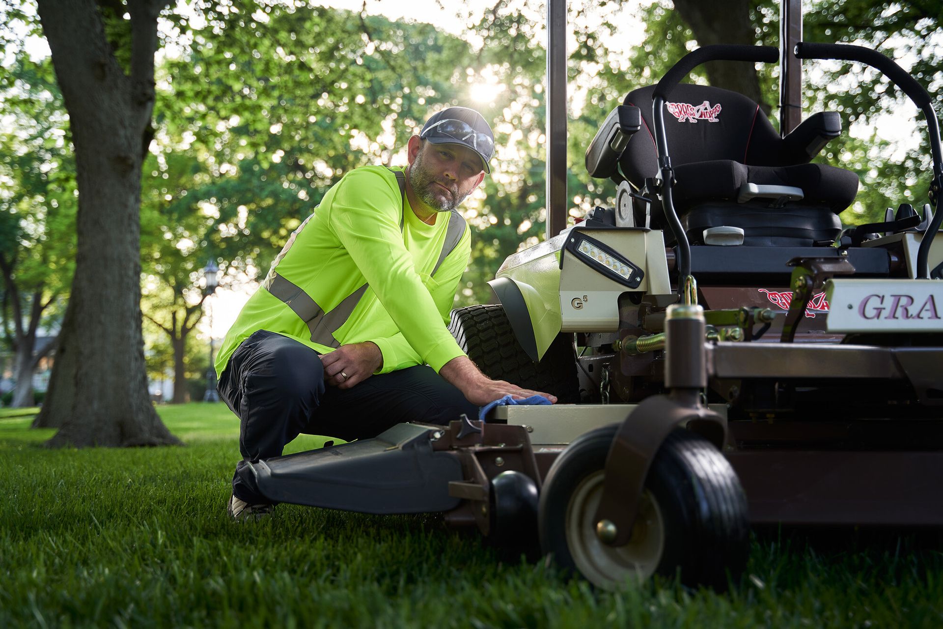 Worker in a hi-vis shirt crouching to wipe a Grasshopper mower's deck with a cloth.