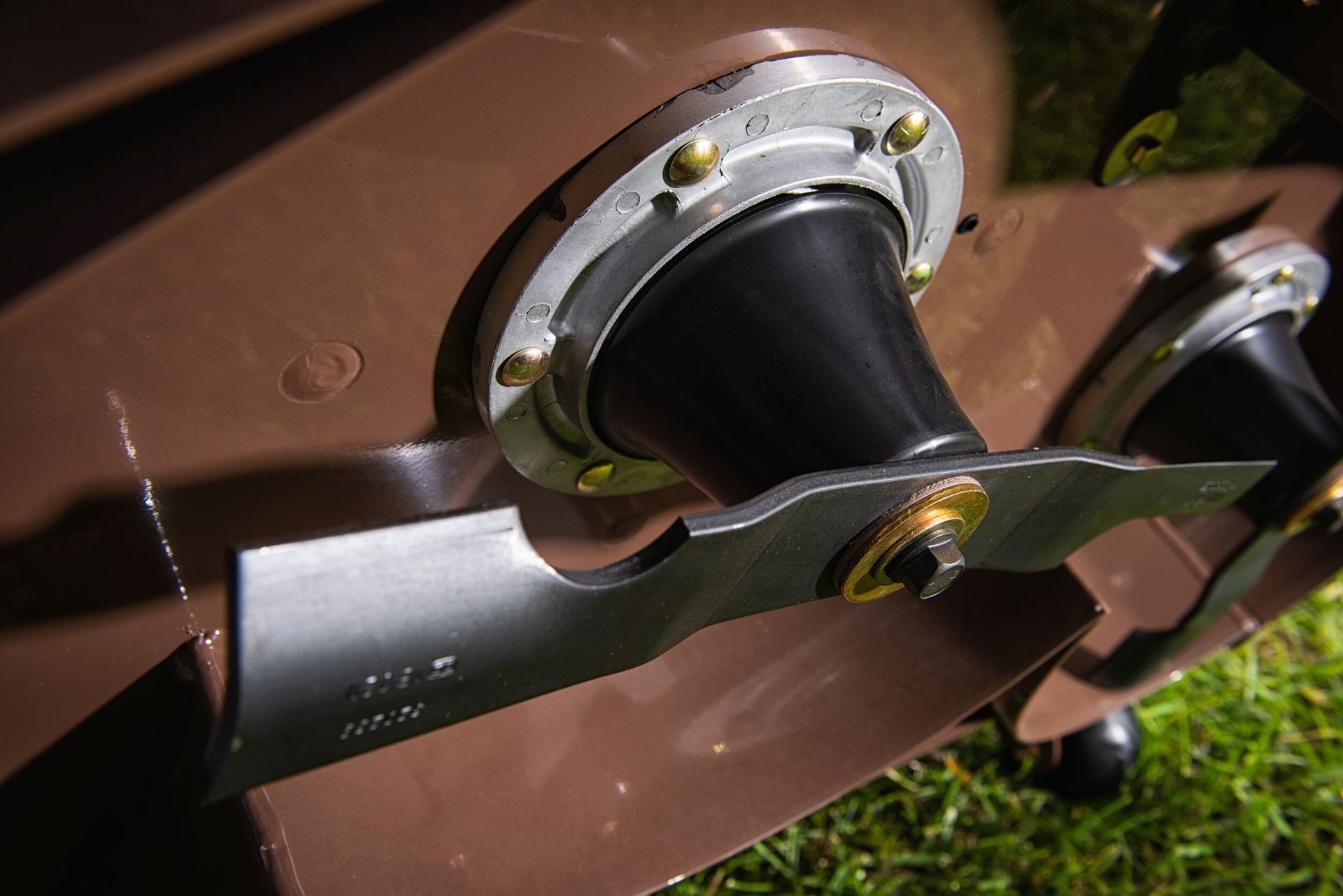 Close-up of a mower deck spindle housing with its bolted blade above green grass.