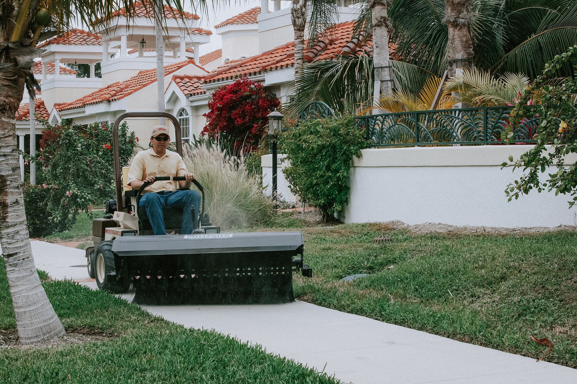 Operator sweeping a concrete walkway with a rotary broom attachment in a palm-lined resort community.