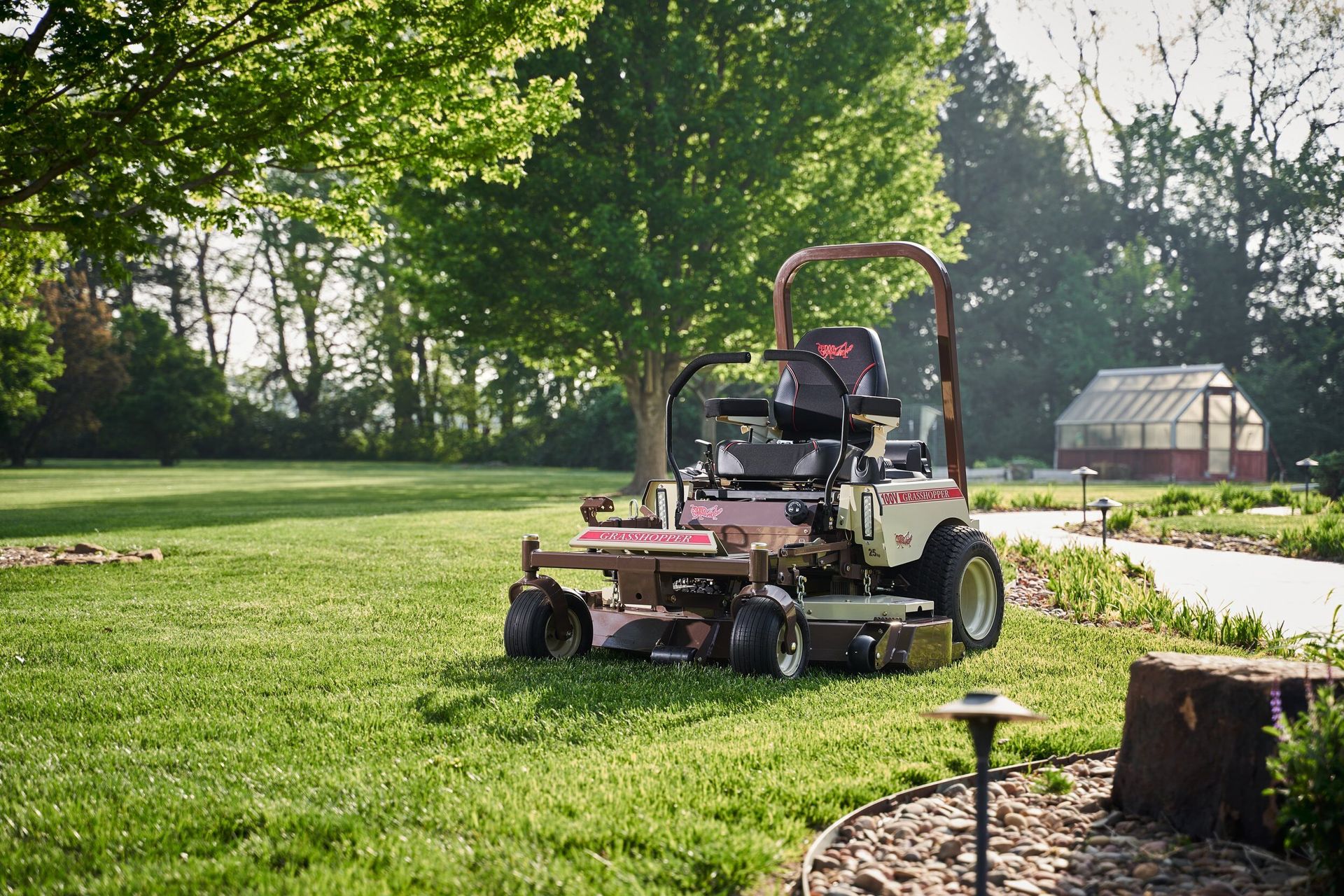 Grasshopper 100V mower parked on a manicured lawn with a backyard greenhouse behind it.