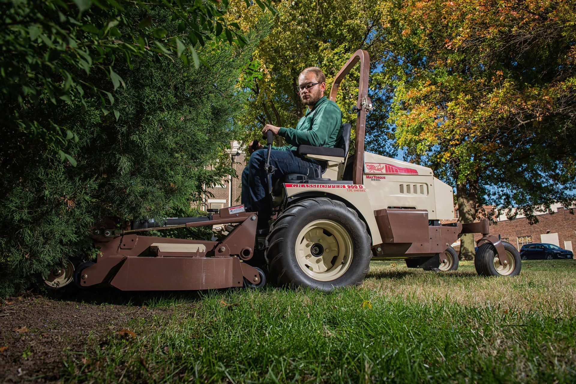 Operator on a Grasshopper 900D diesel FrontMount mower, deck trimming beneath dense evergreen branches.