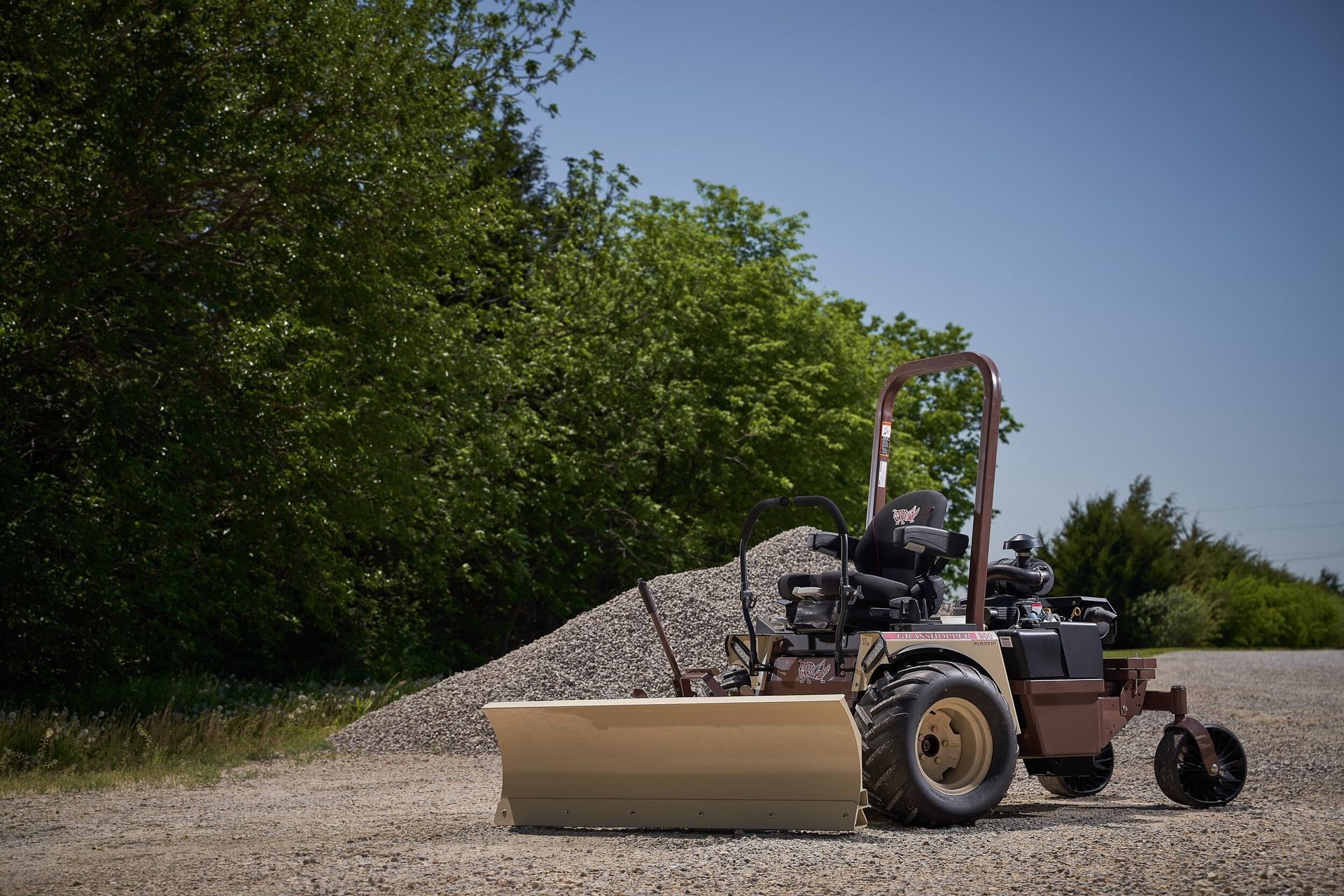 Grasshopper mower fitted with a tan dozer blade, parked on gravel beside a crushed-stone pile.