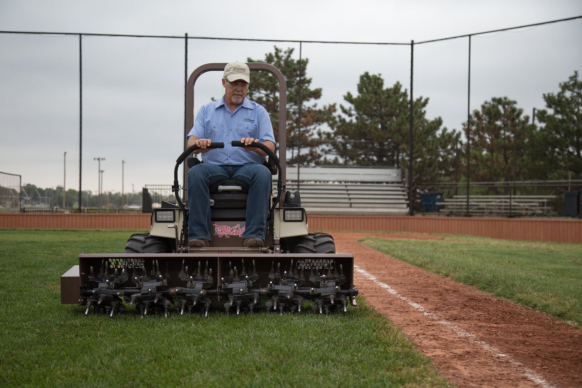 Groundskeeper aerating ballfield turf beside a chalked foul line, with a backstop and bleachers behind.