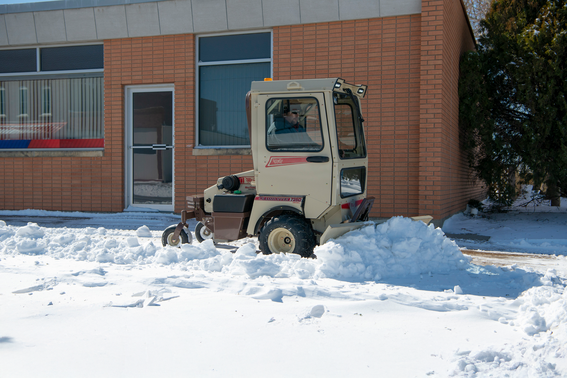 Enclosed-cab Grasshopper 725D diesel pushing a snow ridge along a brick commercial building.