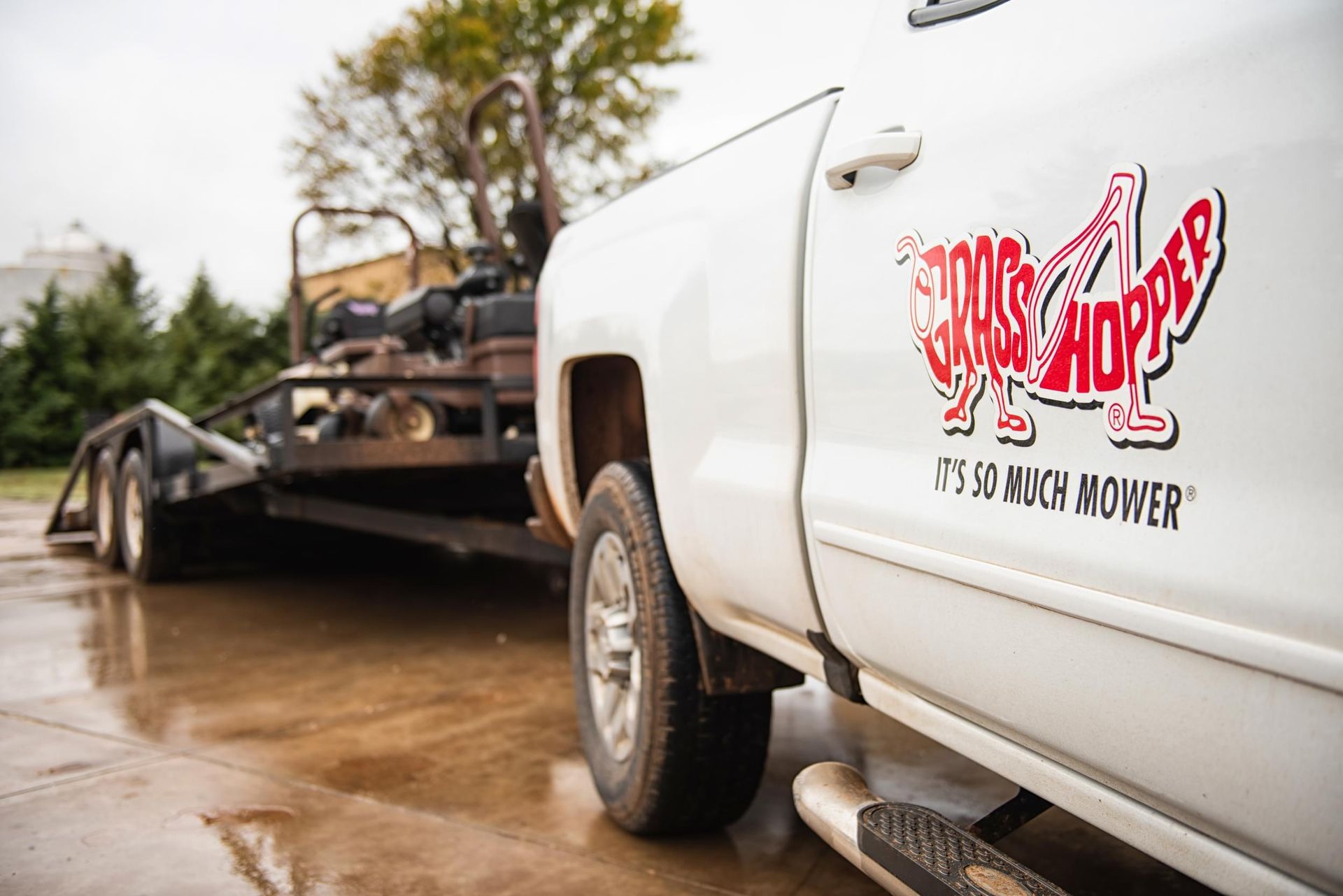 Close-up of a white pickup door bearing the red Grasshopper logo, mower and trailer behind.