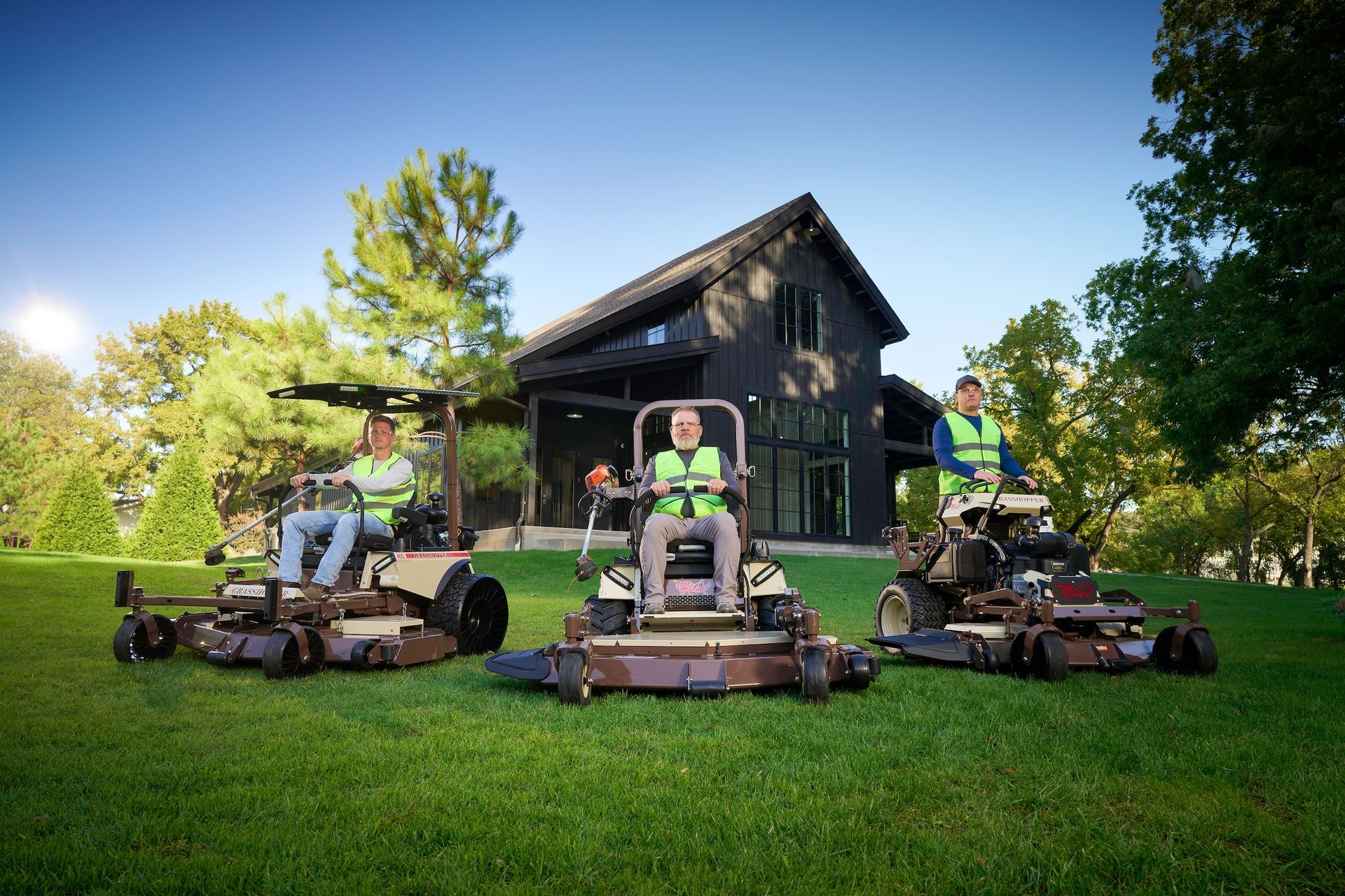 Three-person crew in hi-vis vests on three Grasshopper mowers parked before a dark barn-style house.