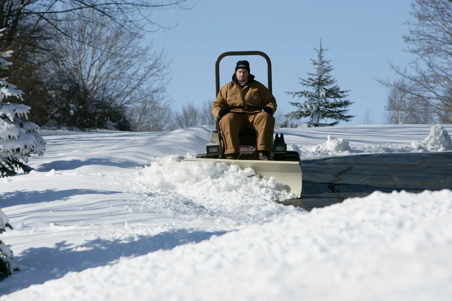 Operator in insulated coveralls pushing snow along a driveway with a mower-mounted dozer blade.