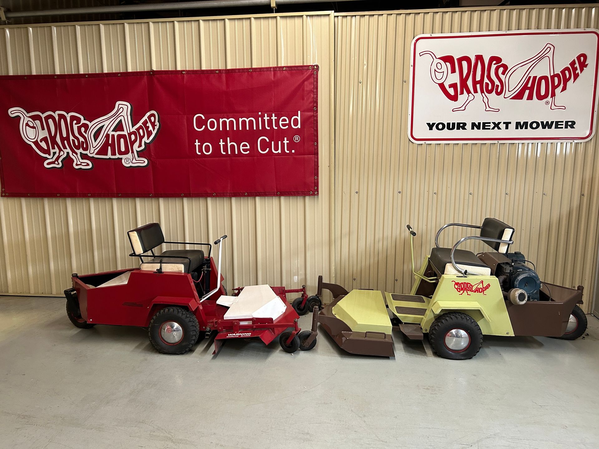 Two vintage Grasshopper mowers, one red and one yellow, displayed indoors beneath Grasshopper banners.