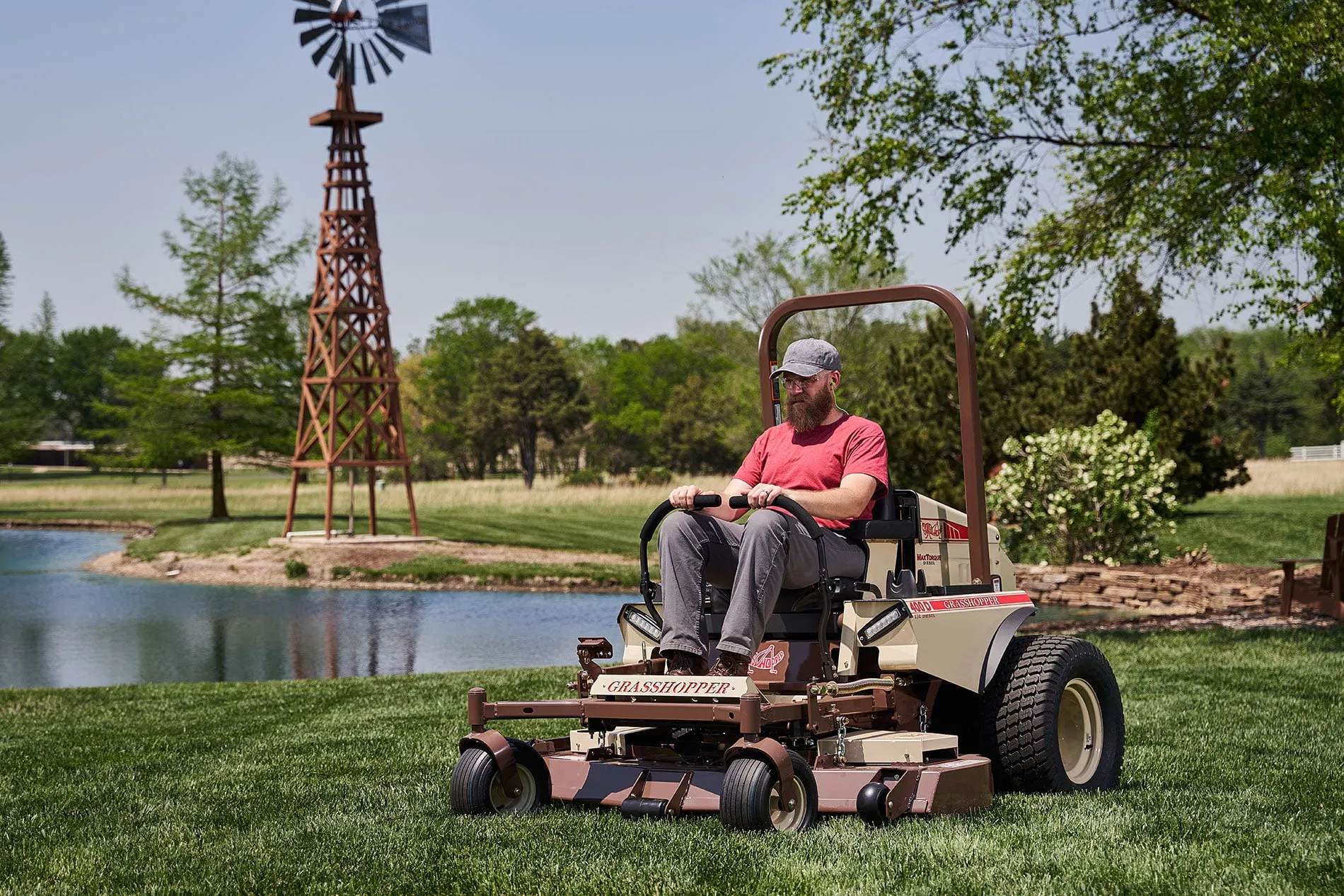 Bearded operator on a Grasshopper 400D diesel FrontMount mower beside a pond and wooden windmill.