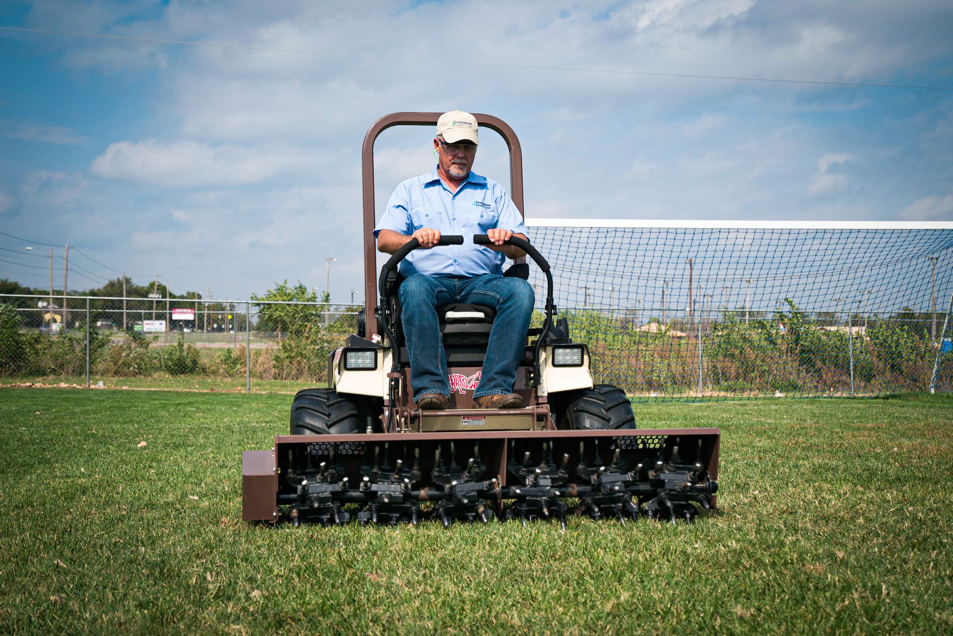 Landscaper operating a Grasshopper mower with an AERA-vator attachment on a sports field.