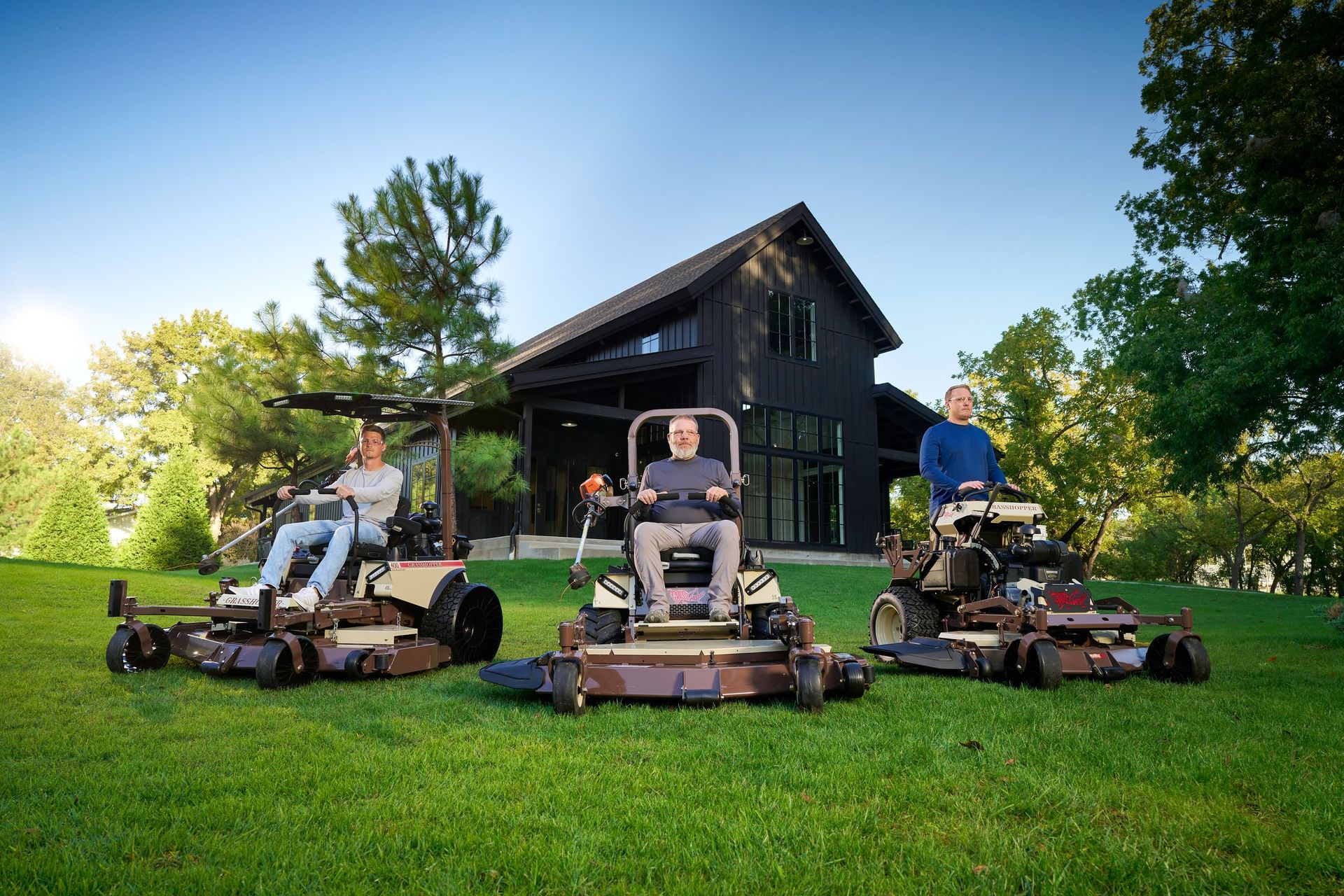 Three operators on seated and stand-on Grasshopper mowers arrayed before a dark barn-style house.