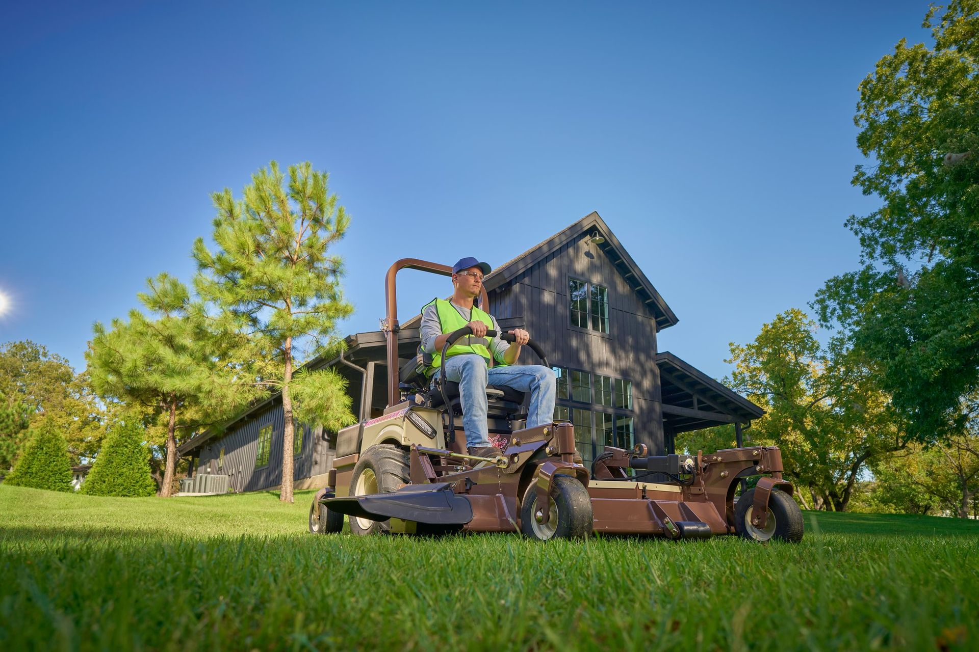 Seated operator mowing a sunlit lawn on a Grasshopper zero-turn mower with an out-front deck.