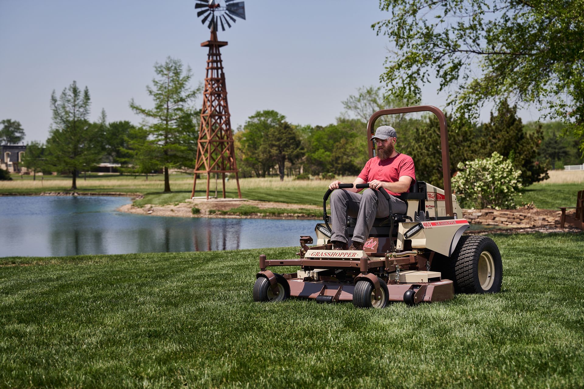 Operator on a Grasshopper 400D diesel mower cutting lawn beside a pond and wooden windmill tower.
