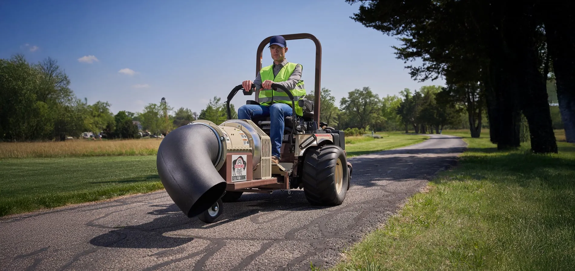 Operator in a safety vest driving a Grasshopper mower fitted with a debris blower along pavement.