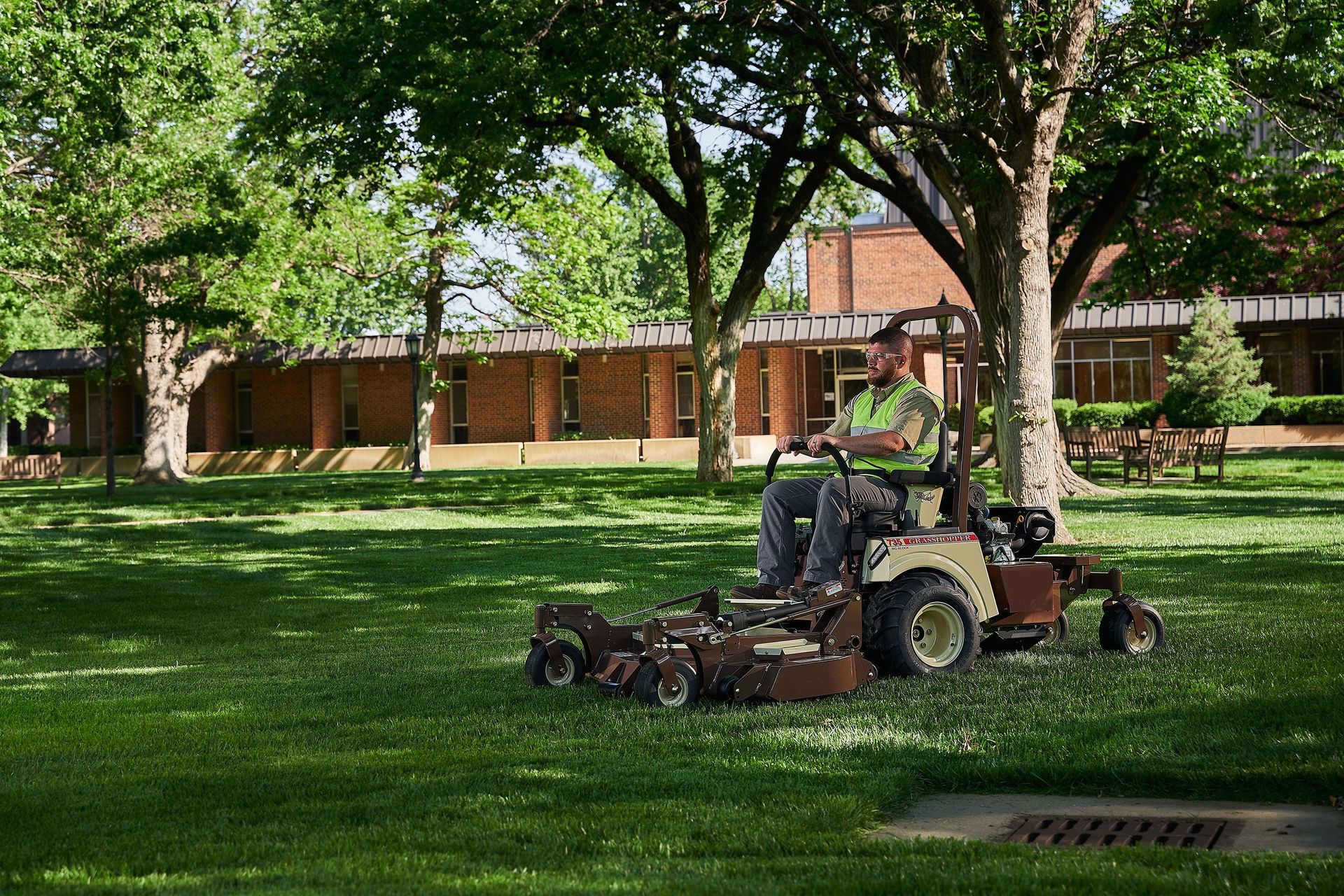 Grounds worker in a hi-vis vest mowing a shaded campus lawn on a Grasshopper 735.
