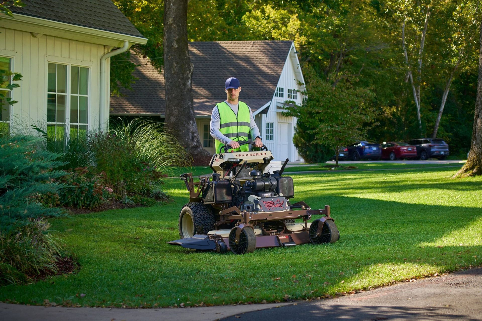 Man in a hi-vis vest operating a Grasshopper stand-on mower across a residential front lawn.