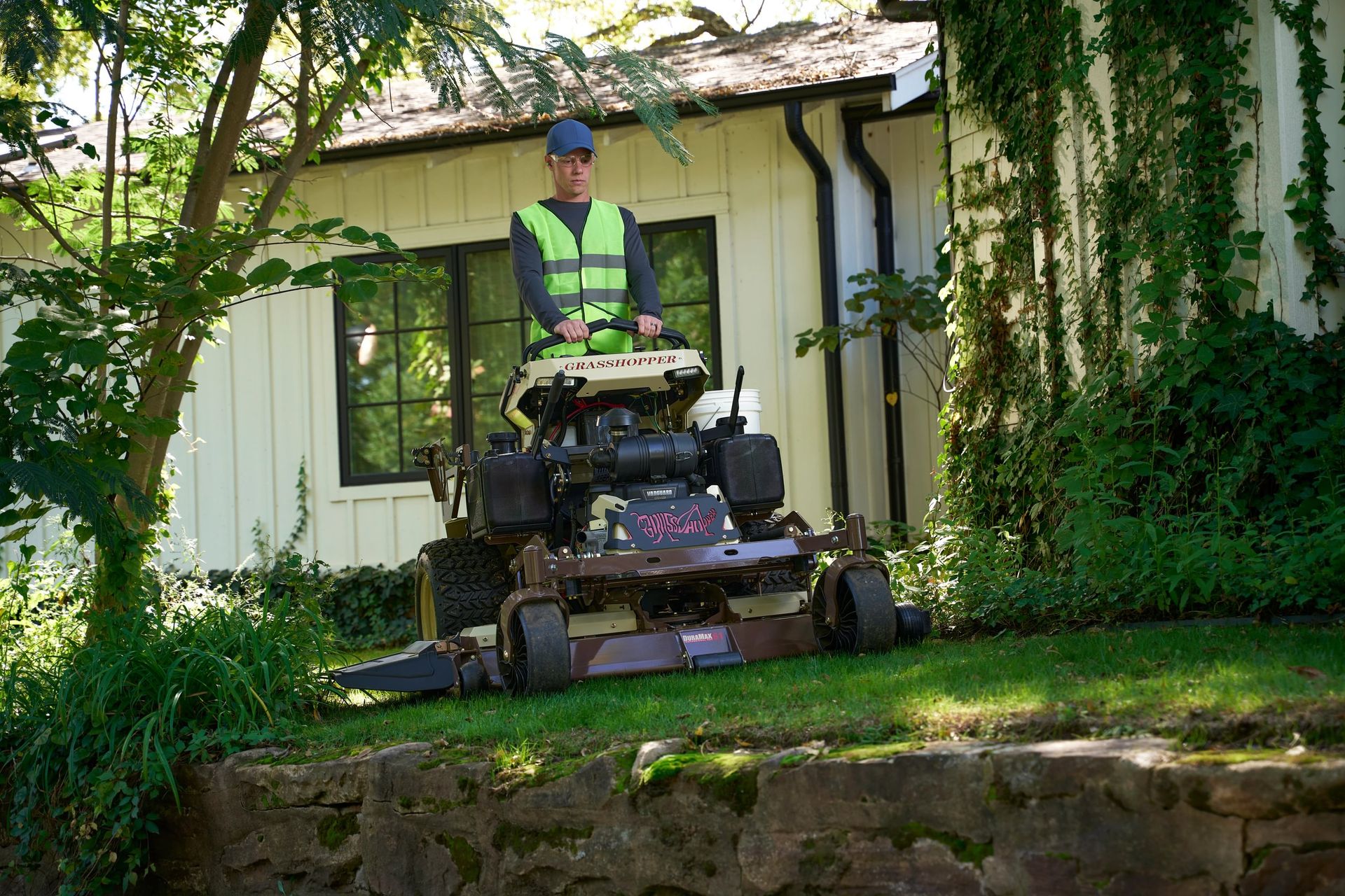 Operator standing on a Grasshopper mower trimming turf beside an ivy-covered house and stone retaining wall.