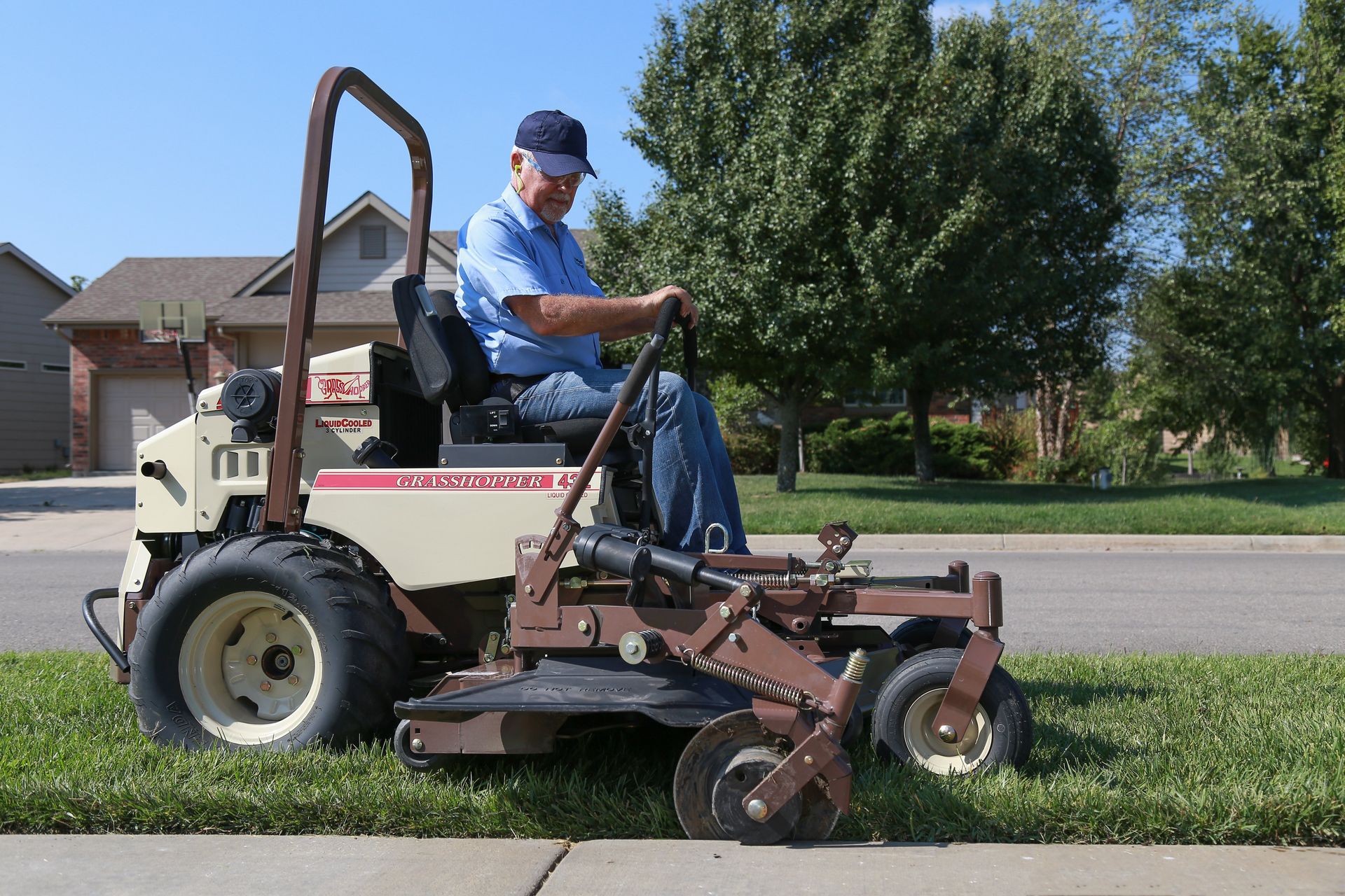Operator on a Grasshopper FrontMount mower cutting a narrow lawn strip beside a residential sidewalk.