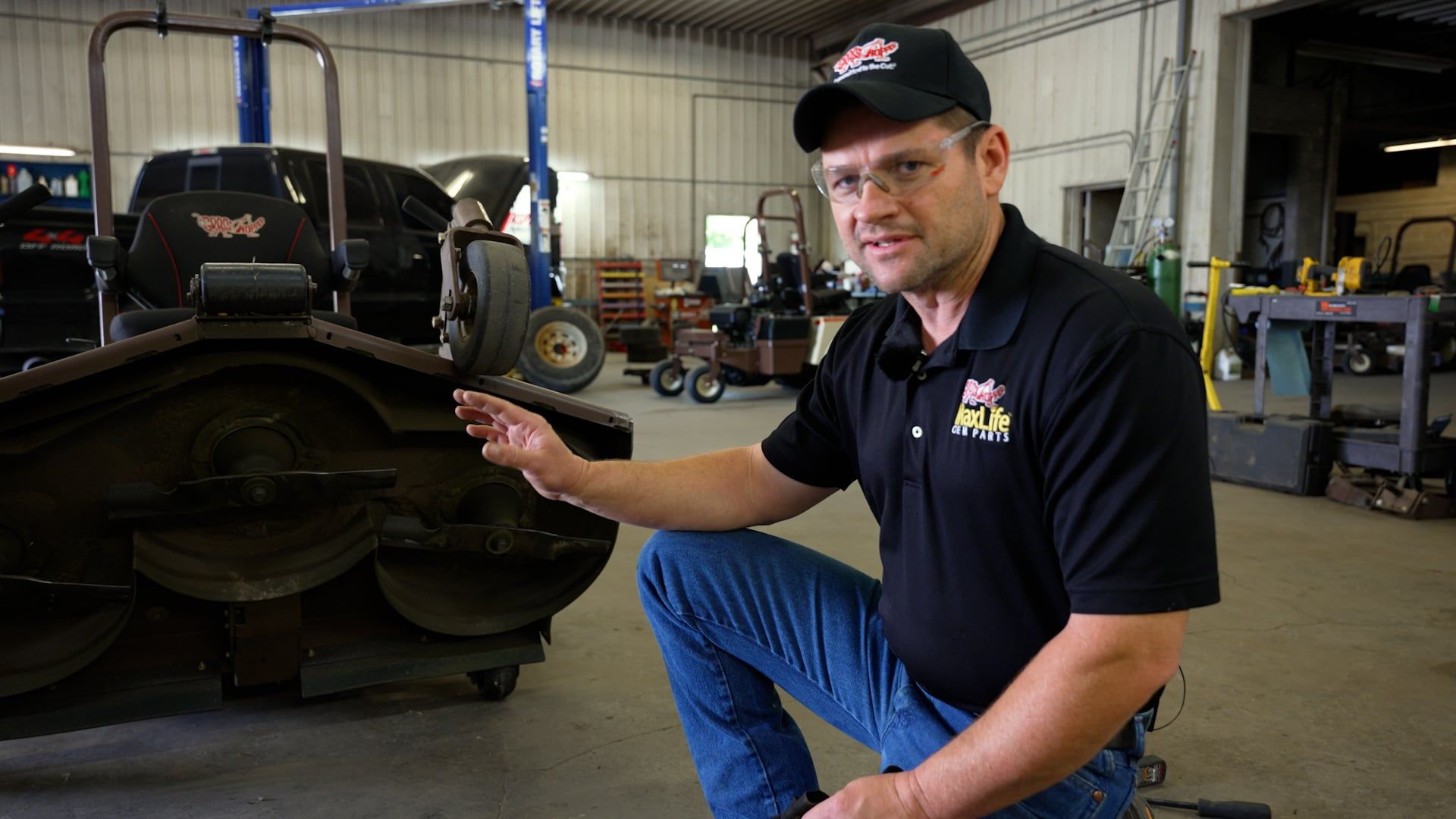 Technician kneeling beside a Grasshopper cutting deck tilted up in a service shop bay.