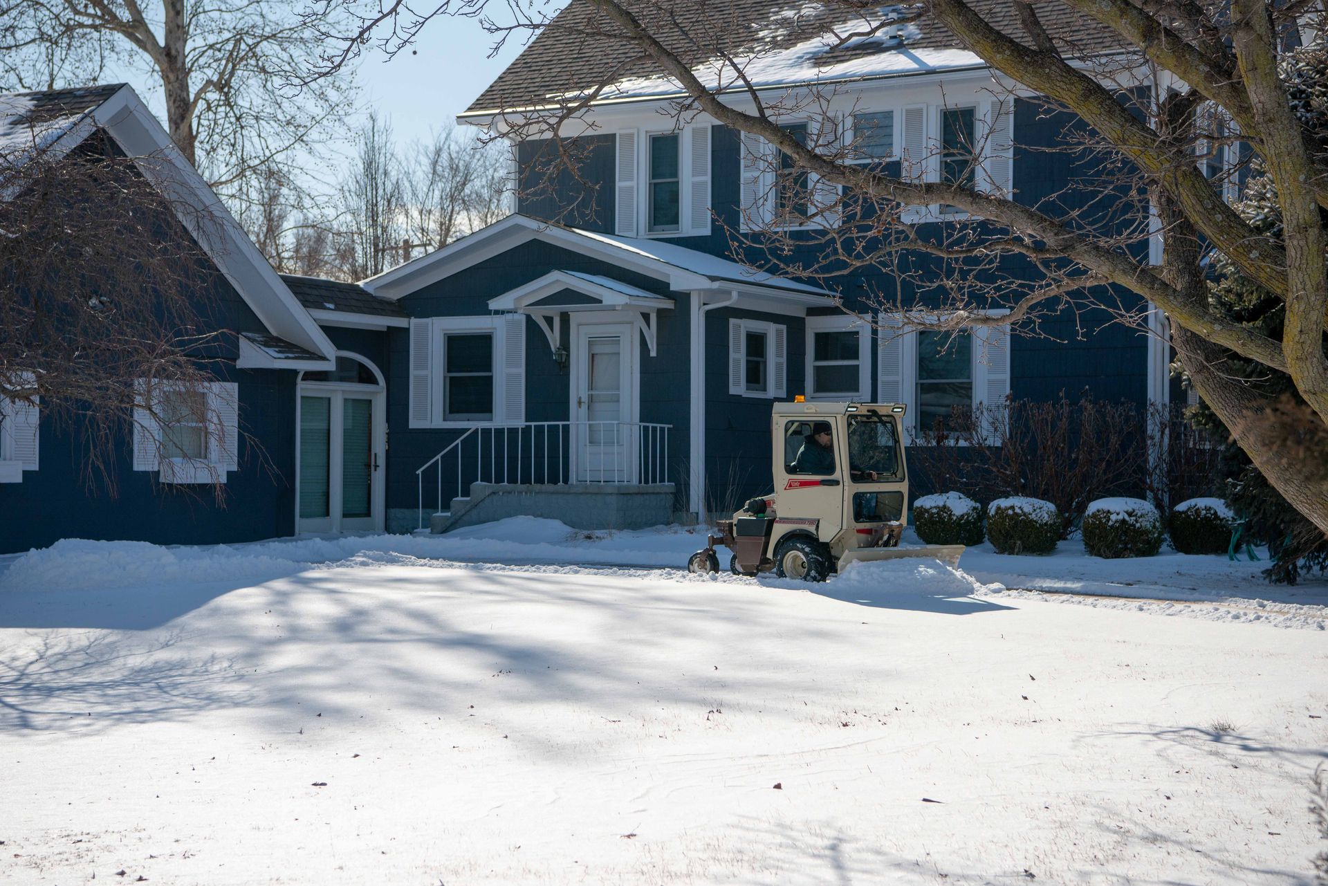 Cab-equipped Grasshopper plowing a snowy driveway in front of a large navy blue house.
