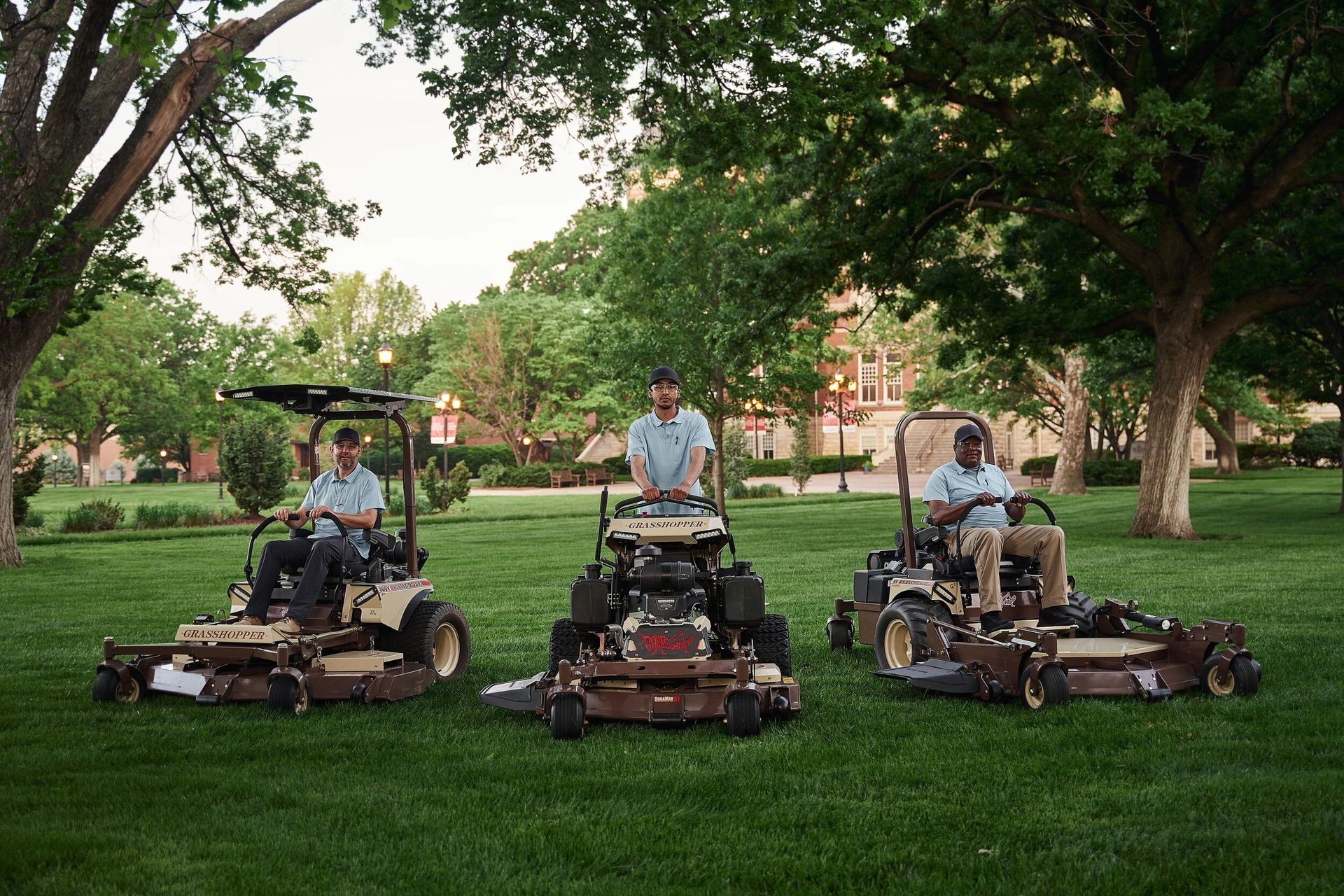 Three grounds crew members with Grasshopper mowers, including a stand-on model, on a campus lawn.