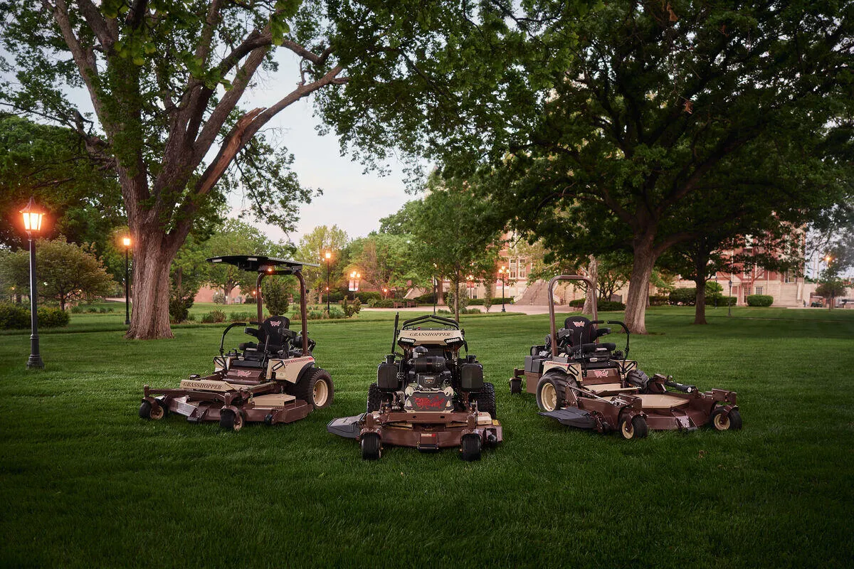 Three Grasshopper mowers, one a stand-on model, parked on a campus lawn at dusk.