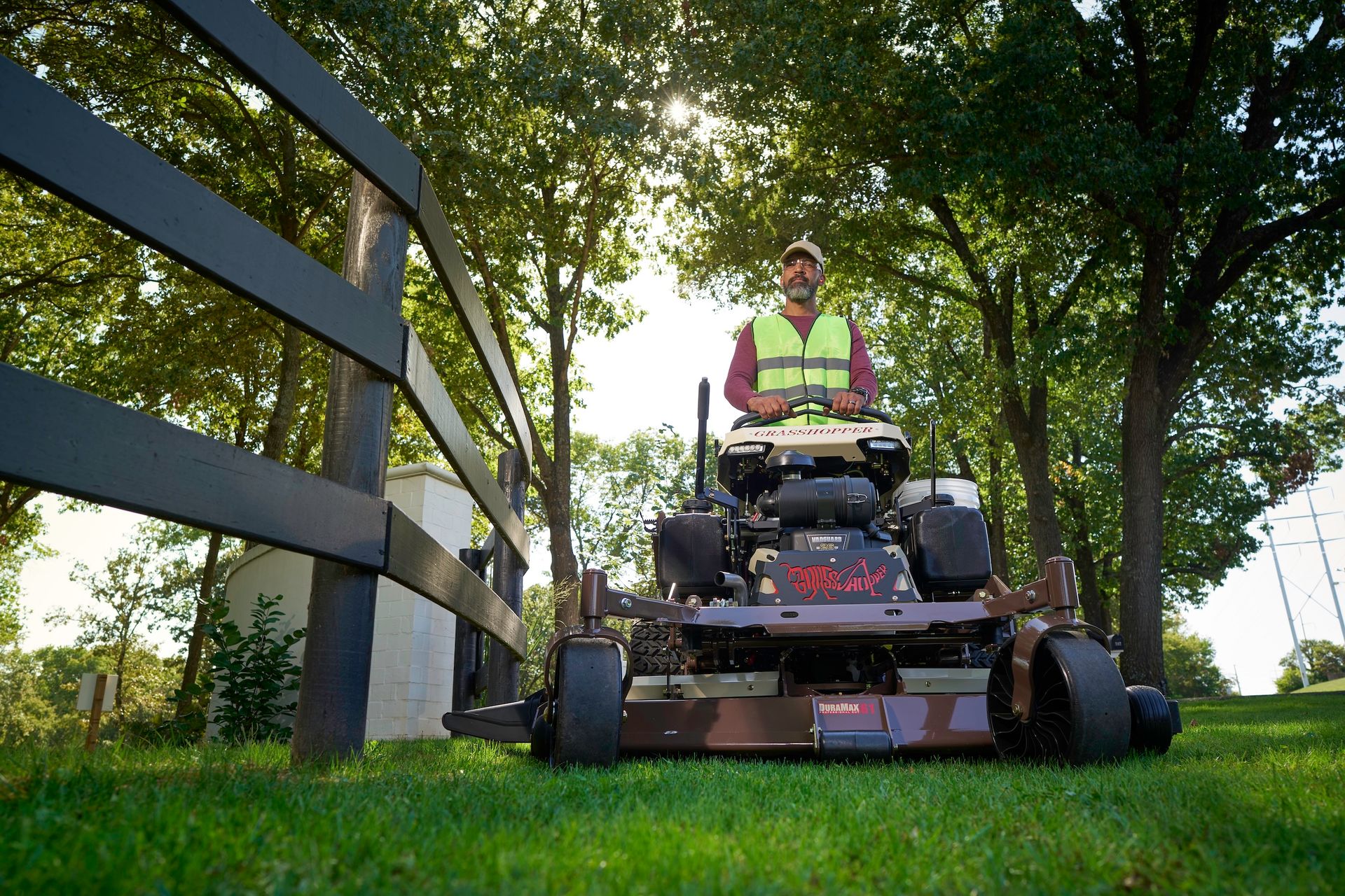 Low-angle view of an operator on a Grasshopper stand-on mower beside a dark rail fence.