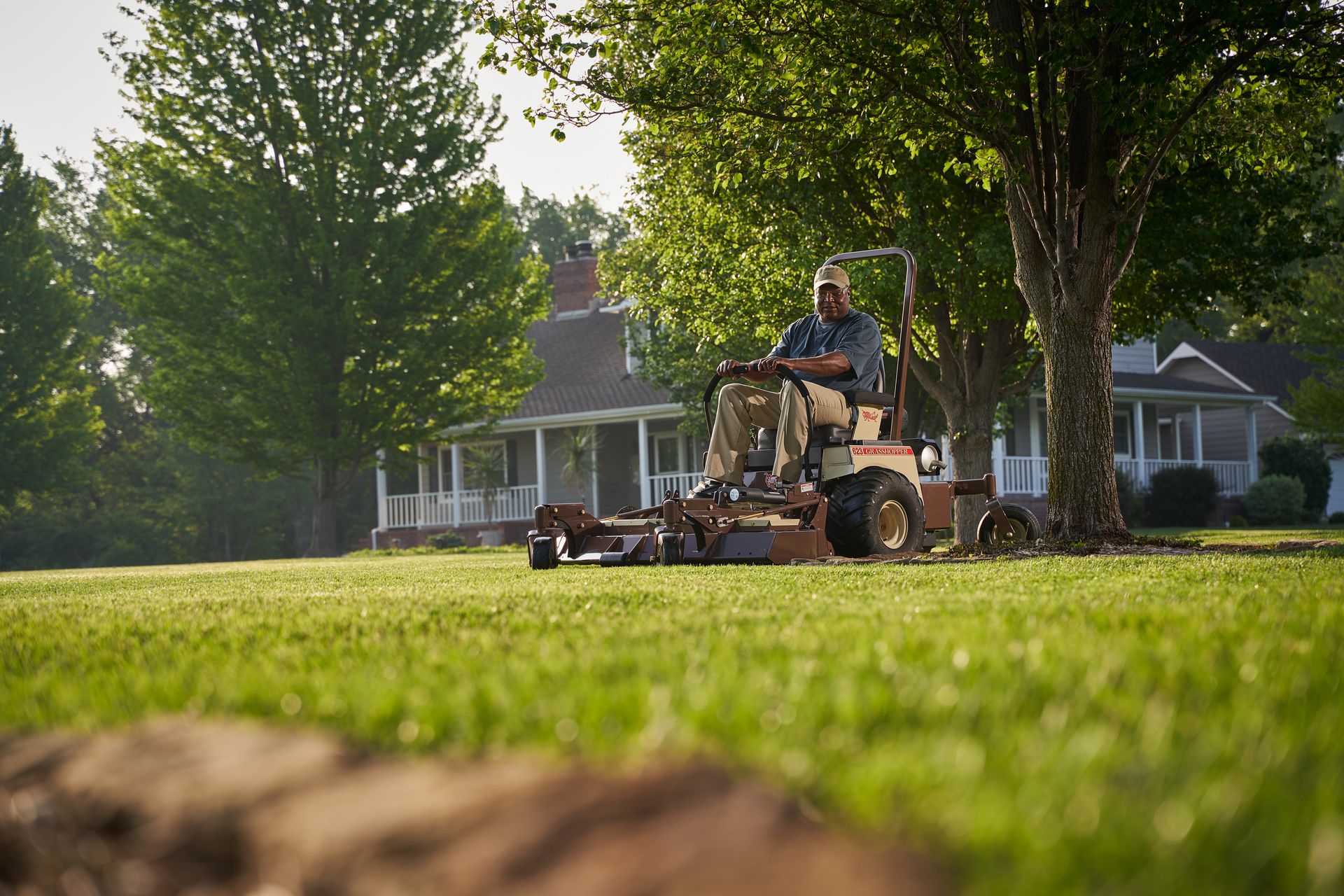 Operator mowing a broad sunlit lawn on a Grasshopper FrontMount mower beside a white-porched house.