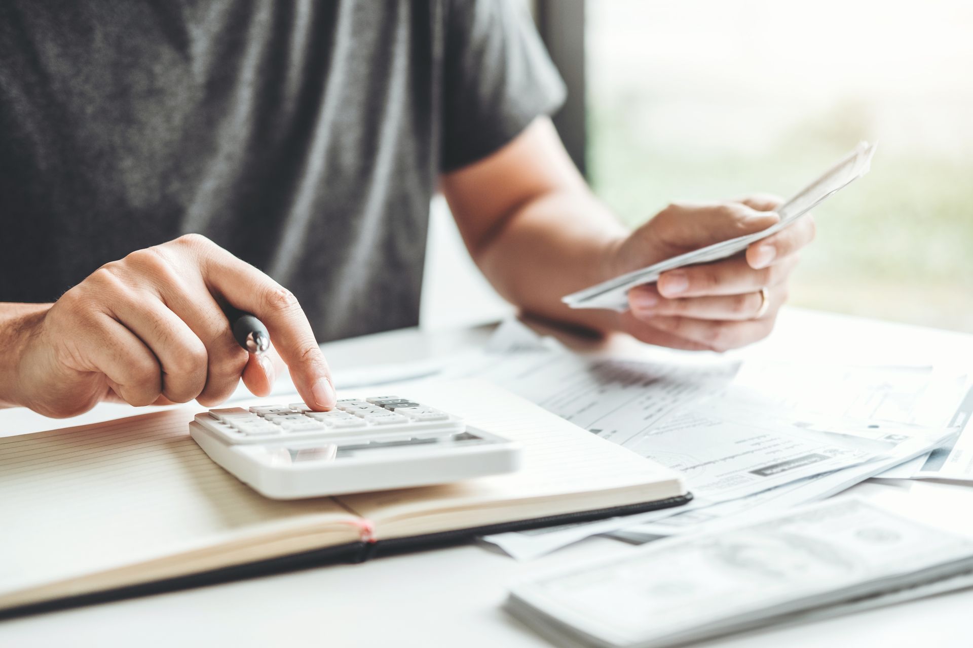 Person tallying paperwork with a calculator beside a notebook, invoices, and US banknotes.