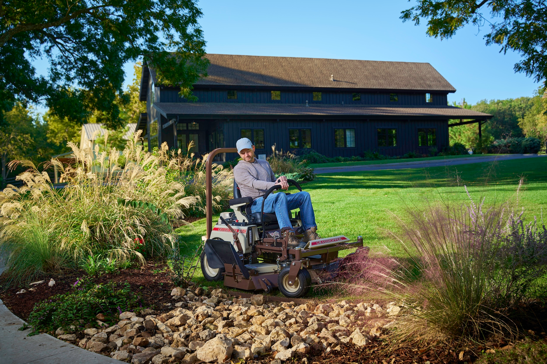 Operator on a Grasshopper 100V mower trimming beside ornamental grasses and a landscaped rock bed.