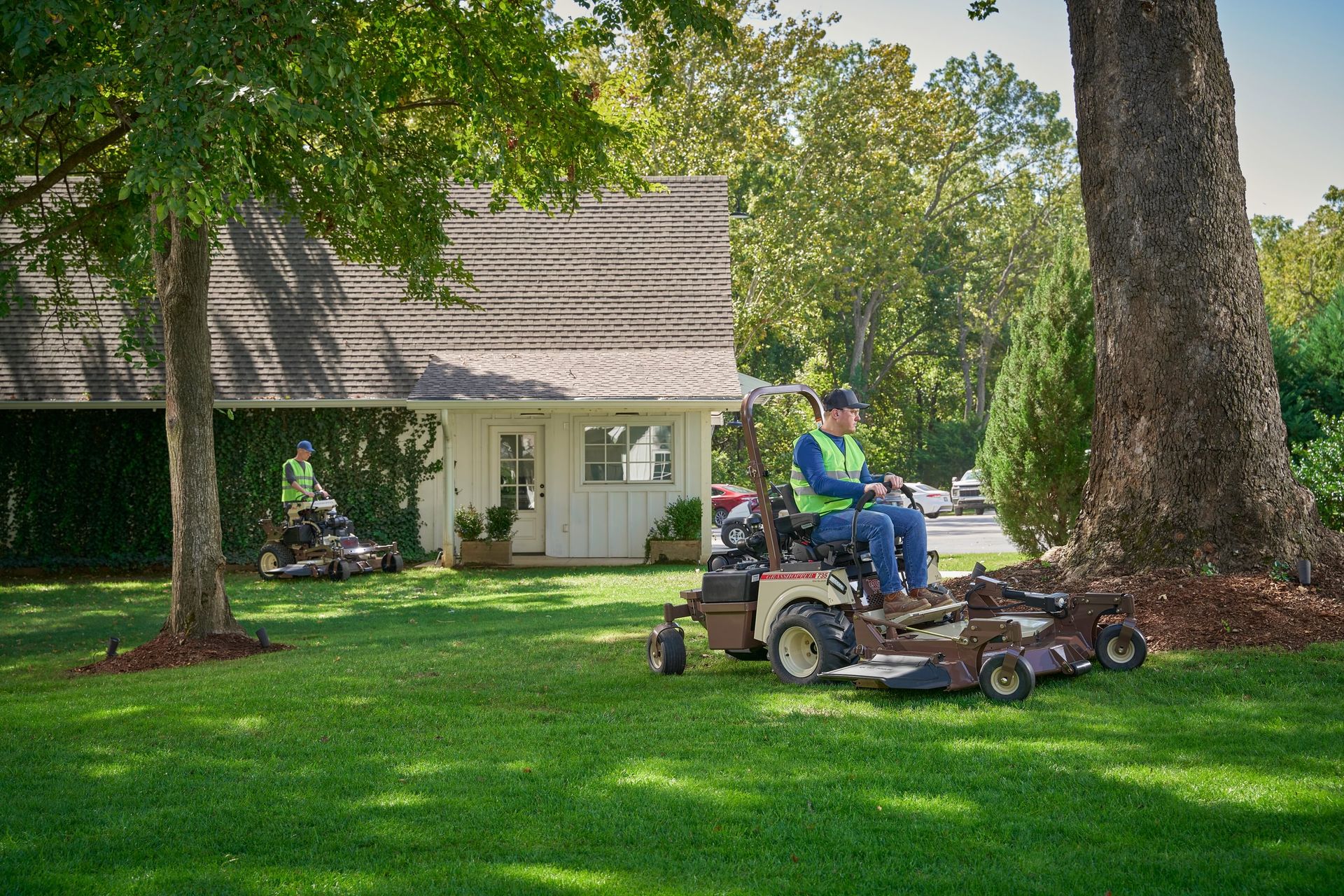 Two operators in hi-vis vests mowing a shaded lawn, the nearer on a Grasshopper 735.