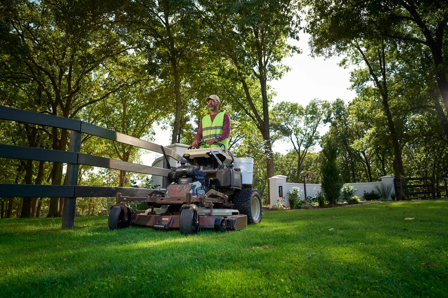 Operator steering a Grasshopper stand-on mower across sunlit lawn between a fence and white wall.