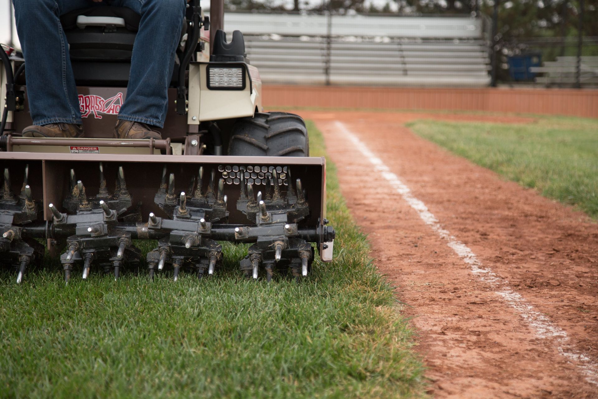 Close-up of a spiked aerator attachment on a Grasshopper mower working turf beside a baseball infield.