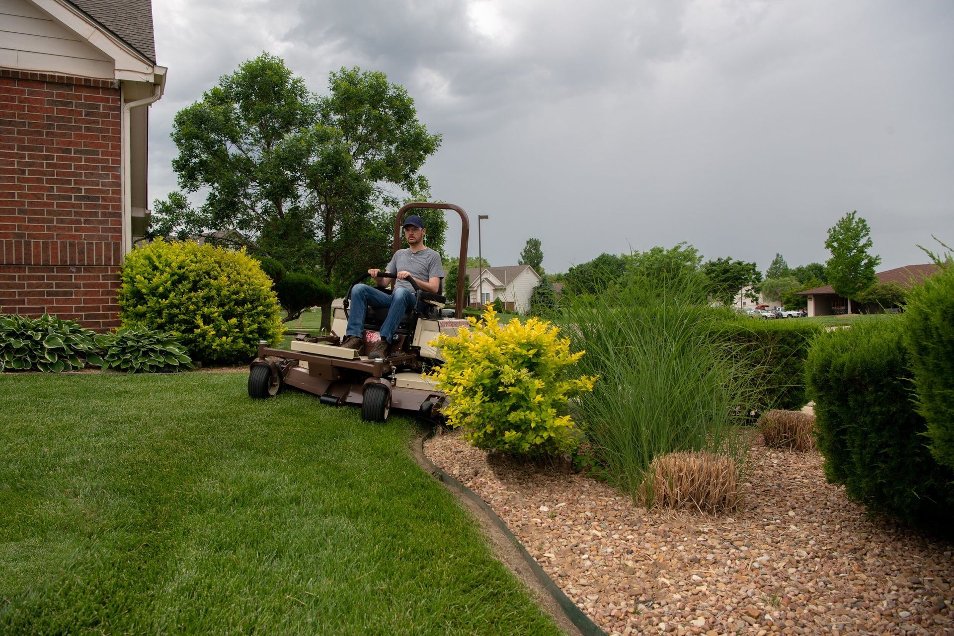 Operator mowing beside gravel landscape beds on a Grasshopper 225V under an overcast sky.