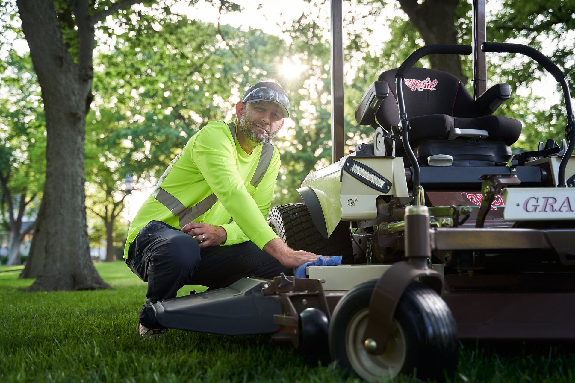Worker in a hi-vis shirt kneeling to wipe down a Grasshopper zero-turn mower on turf.