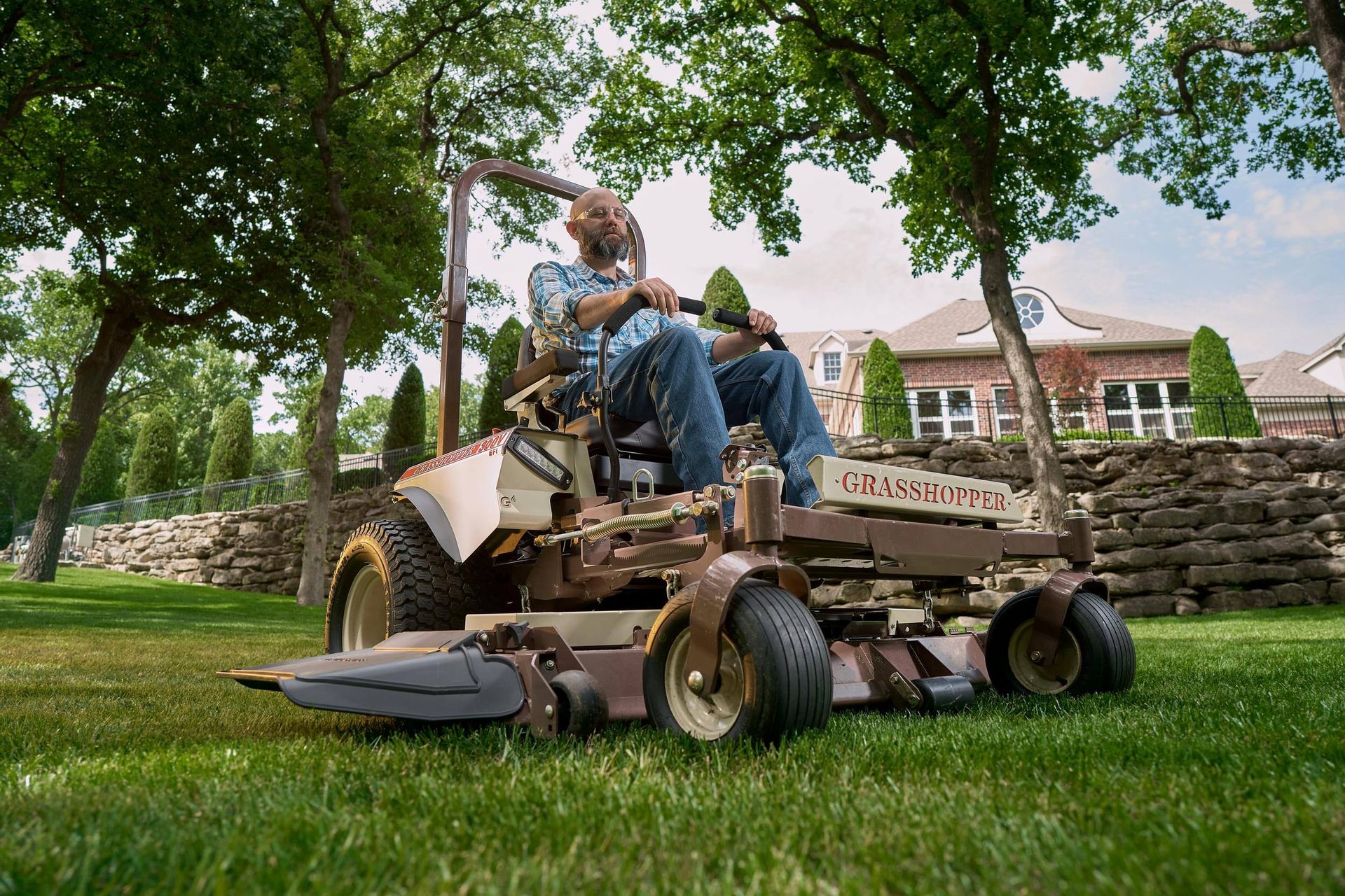 Low-angle shot of a man on a Grasshopper 300V mowing lawn beside a stone retaining wall.