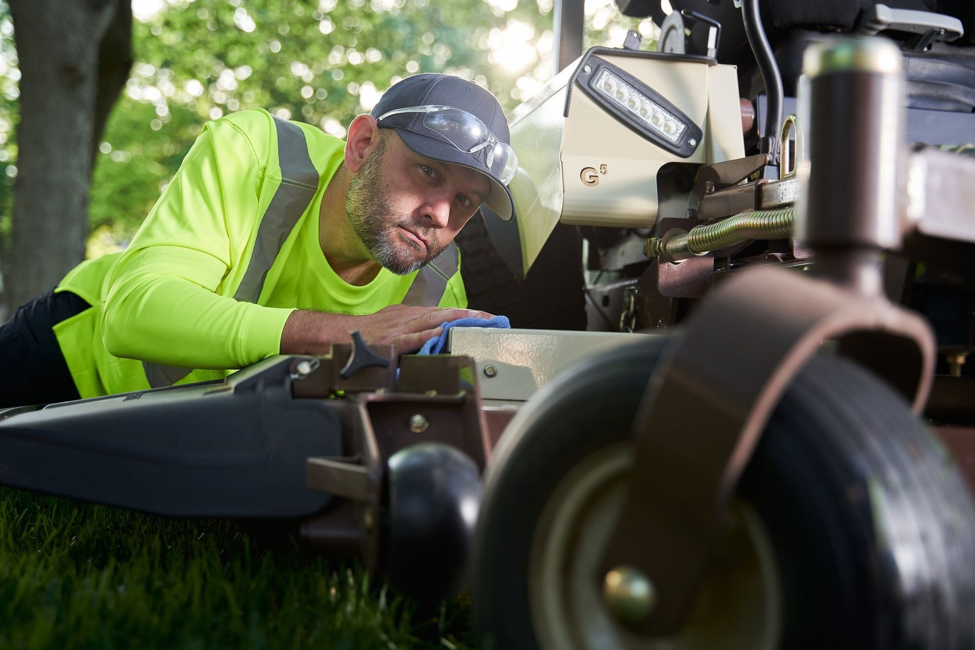 Worker in a hi-vis shirt lying on turf wiping a cutting deck with a blue cloth.