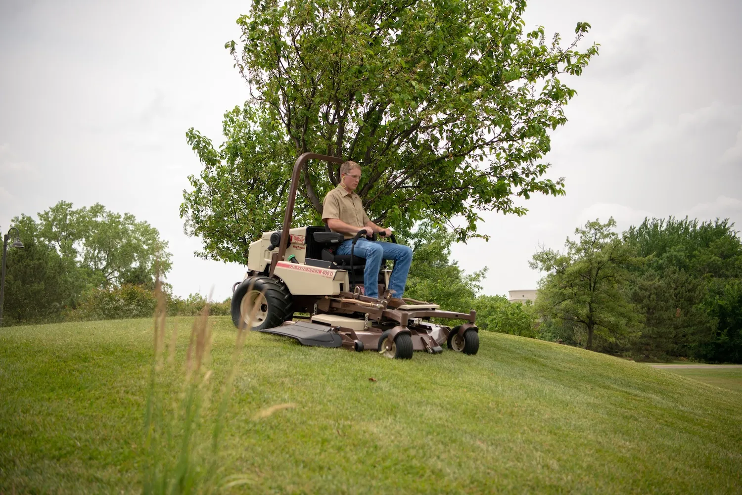 Operator in safety glasses mowing a grassy slope on a Grasshopper 400D beneath a small tree.
