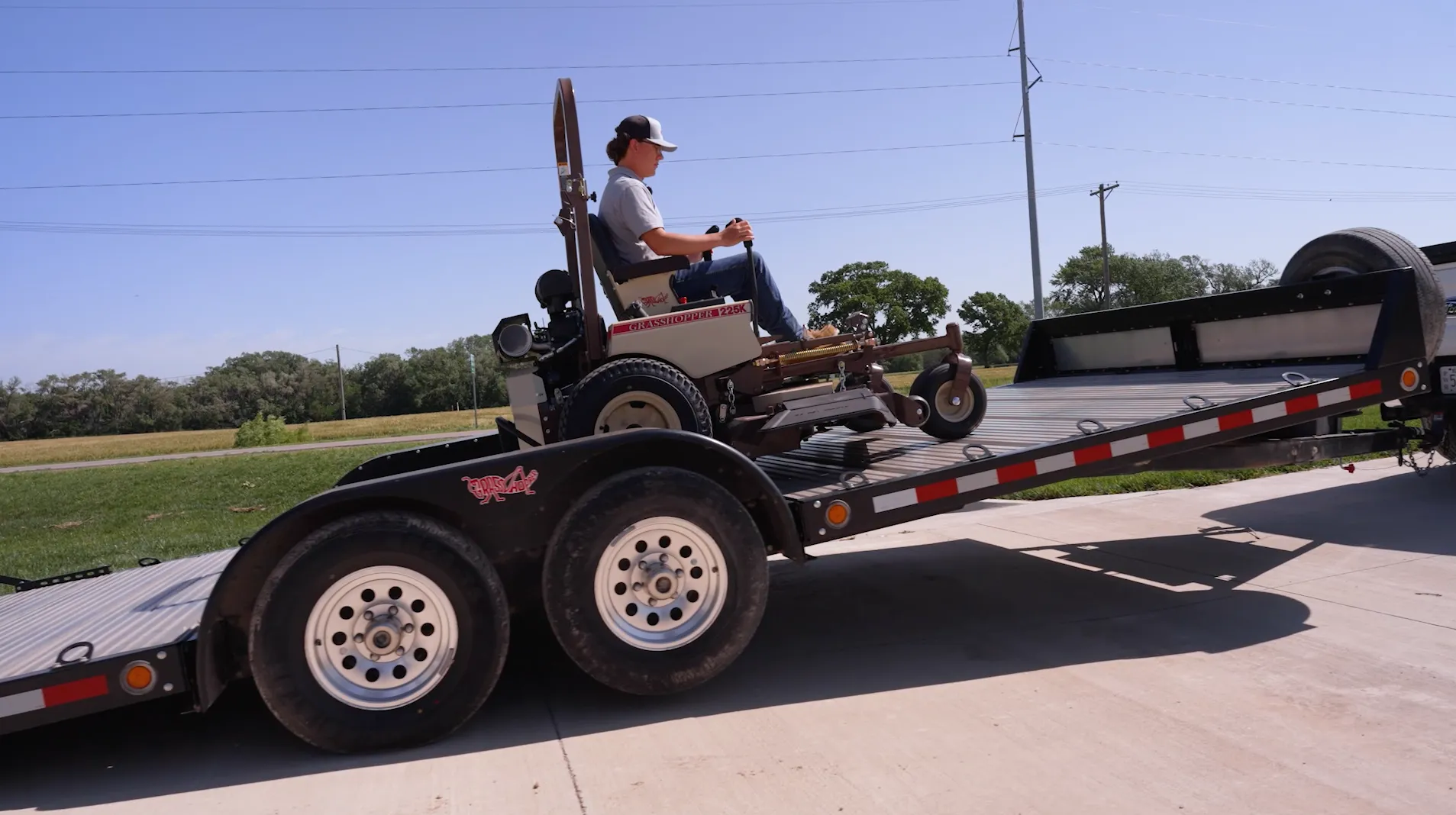 Operator driving a Grasshopper 225K mower down the tilting ramp of a black trailer.
